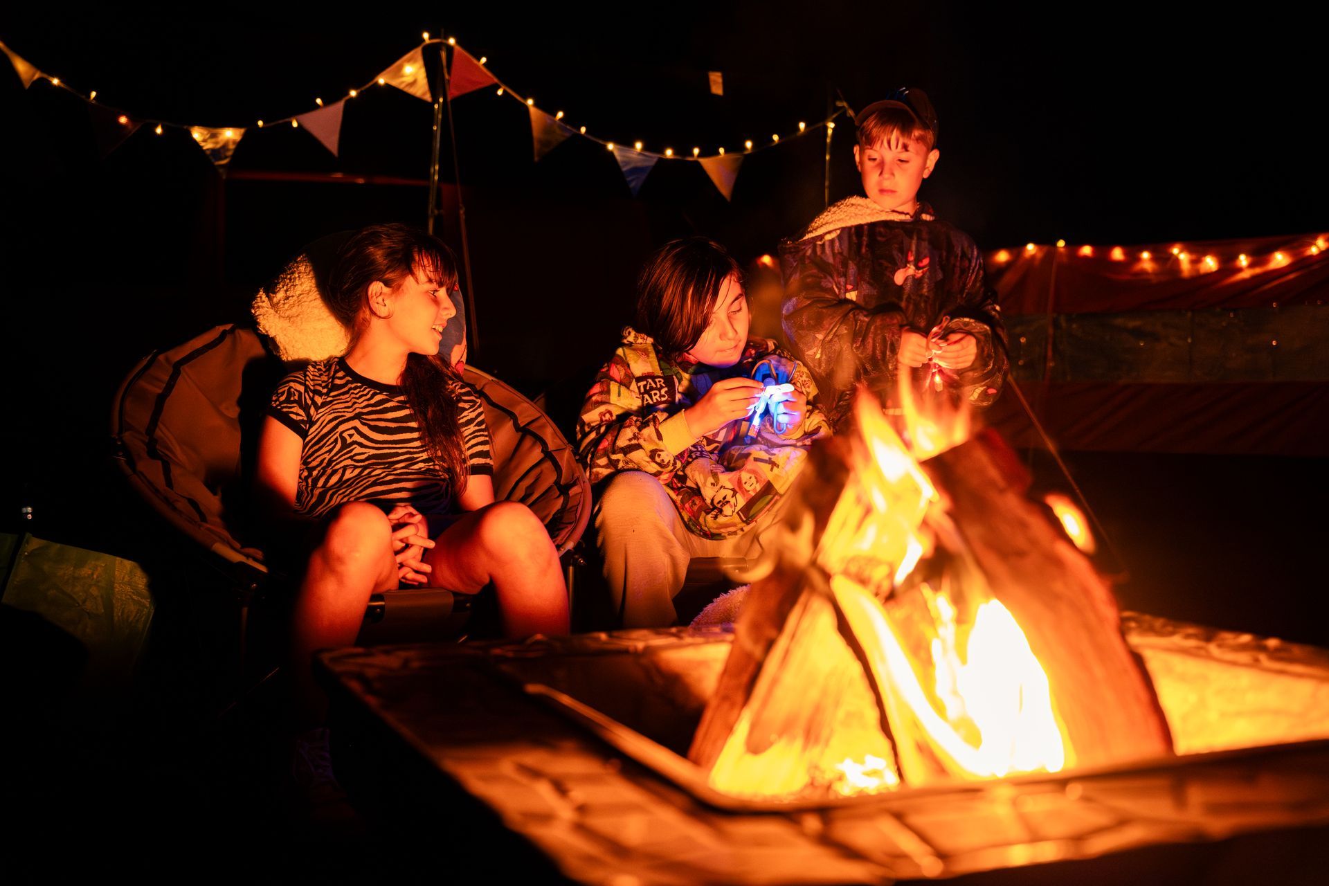 Three people around a campfire at night. String lights and flags hang above.