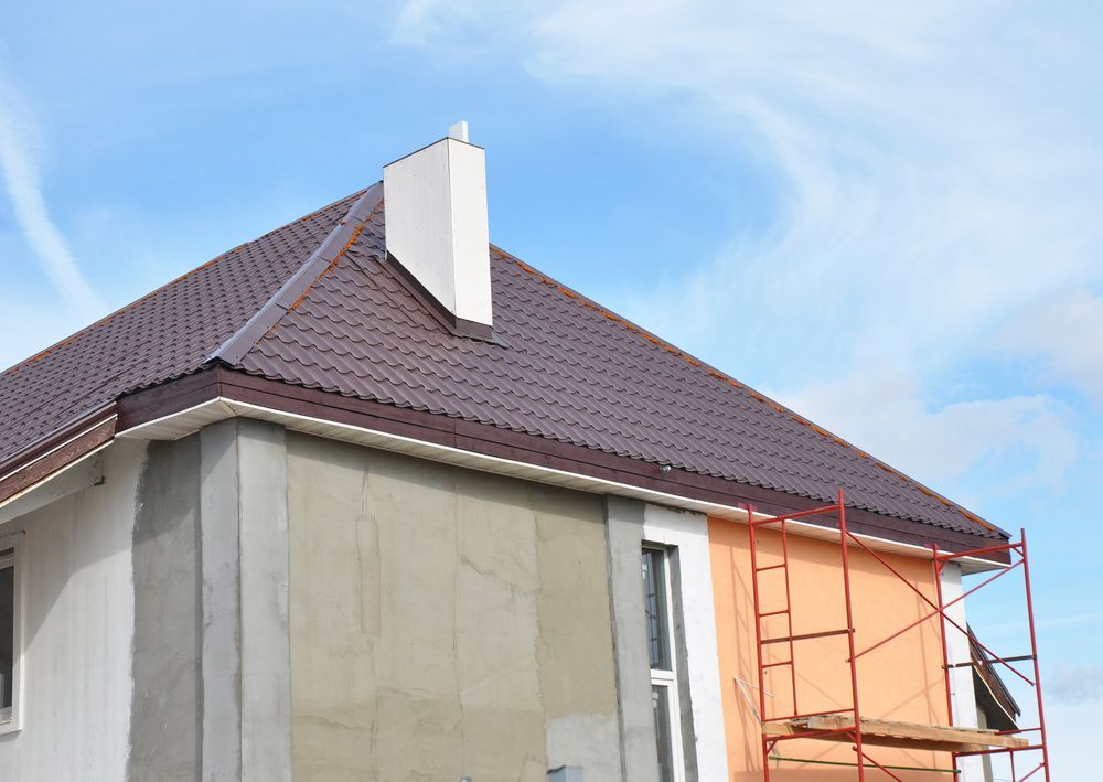 Casa in costruzione con tetto marrone, camino bianco e impalcature arancioni contro un cielo azzurro.