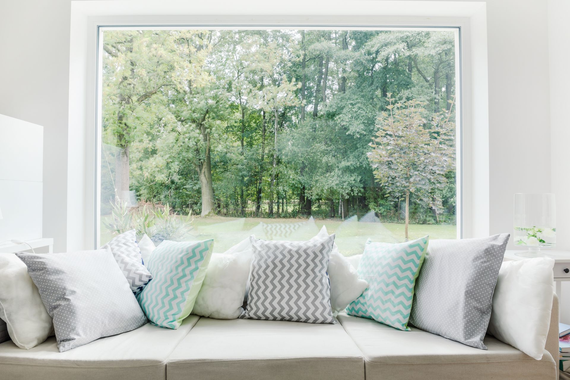 Bright living room with a beige sofa, patterned pillows, and a large window overlooking green trees