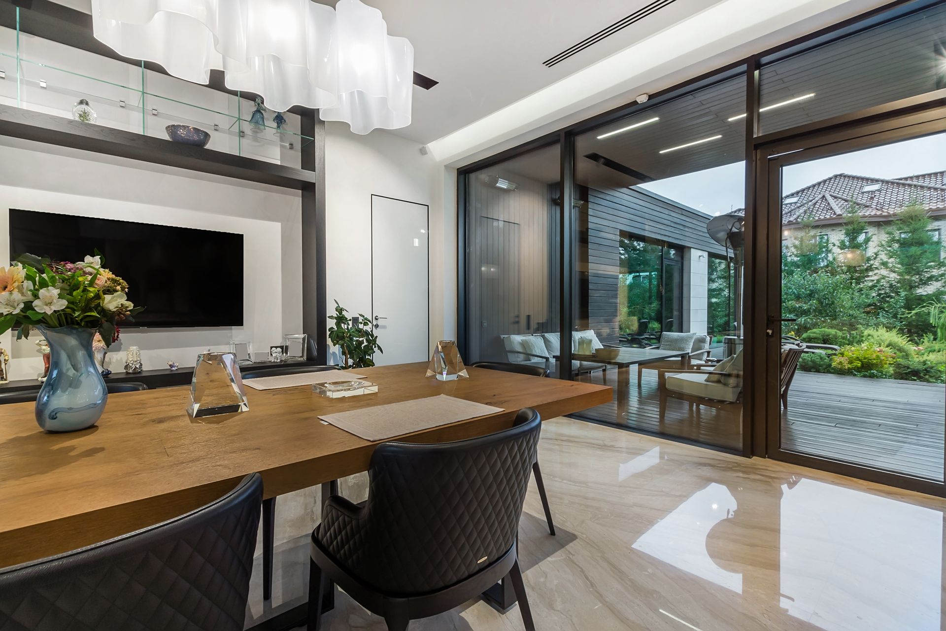 Modern dining room with wooden table, black chairs, and floor-to-ceiling glass doors opening to a garden.