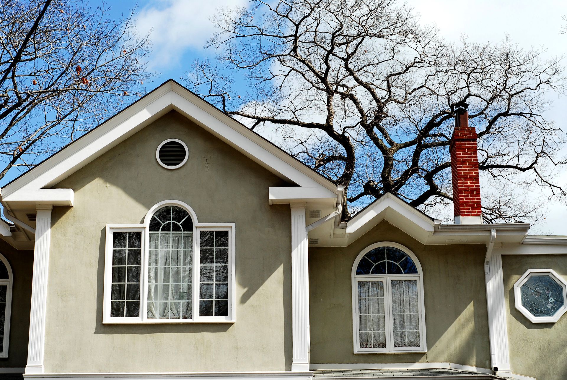 Suburban house exterior with beige walls, arched windows, and a red brick chimney under leafless trees