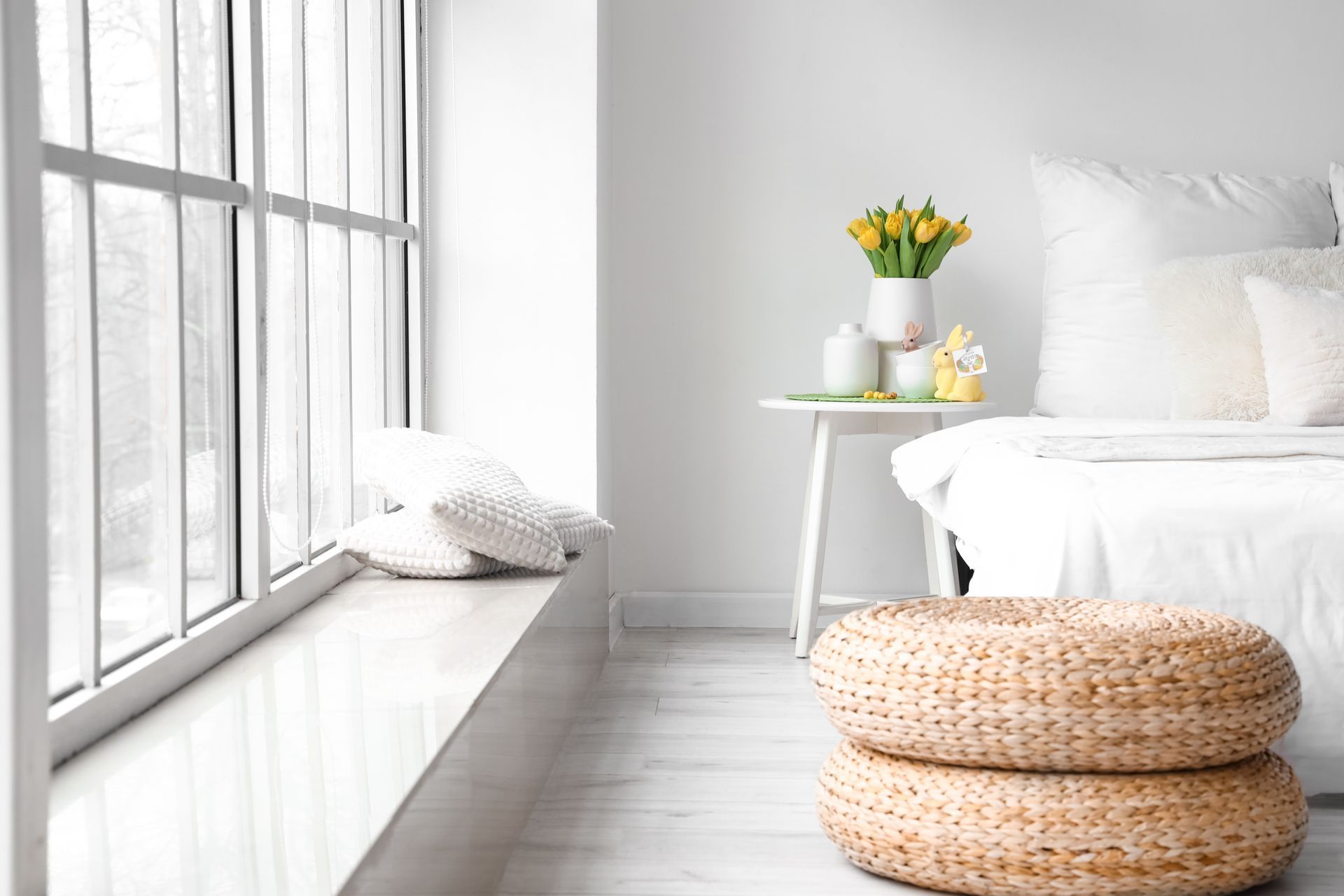 Bright white bedroom with a window seat, bed, flowers on a side table, and woven poufs