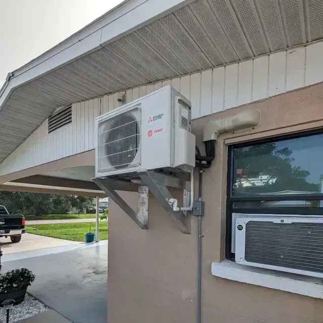 Hvac-Contractor-Bradenton-White mini-split air conditioner mounted on the exterior wall of a beige house next to a window.