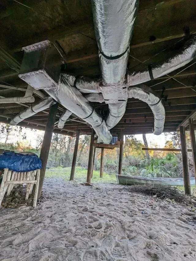 Hvac-Contractor-Bradenton-Insulated air ducts snake across the underside of a wooden structure. Sandy ground, supporting posts, trees in the background.