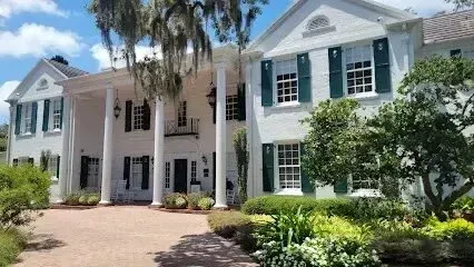 Hvac-Contractor-Bradenton-A large white house with green shutters is surrounded by trees and bushes.