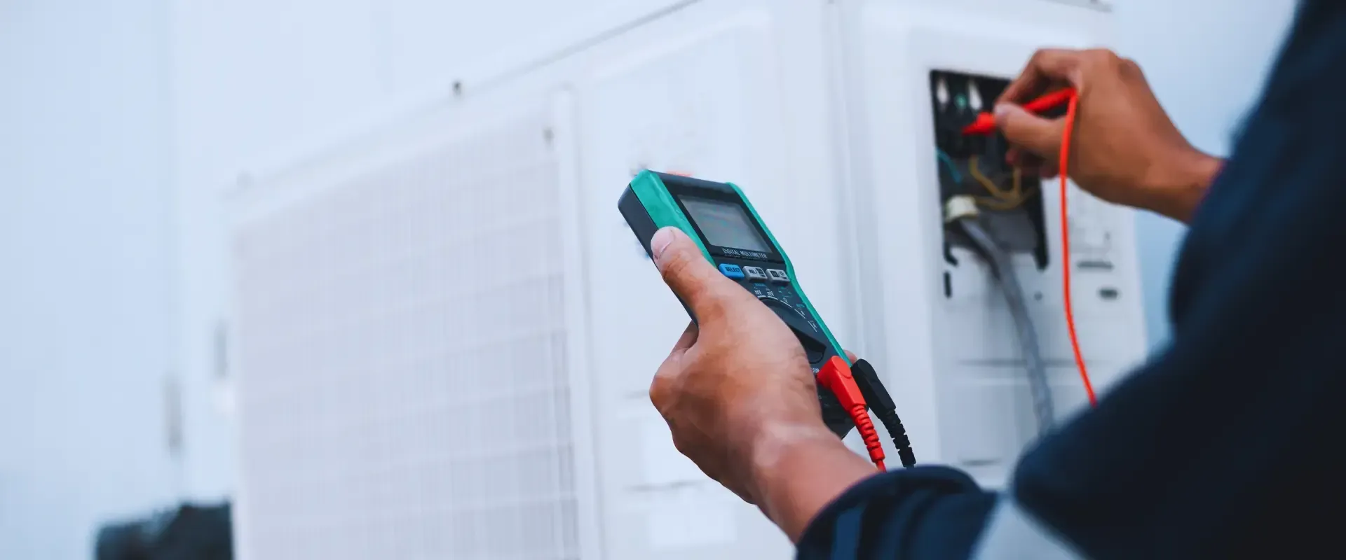 A man is holding a multimeter in his hand while working on an air conditioner.