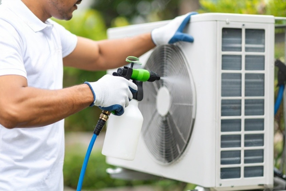 Man wearing gloves sprays a white air conditioning unit outdoors.