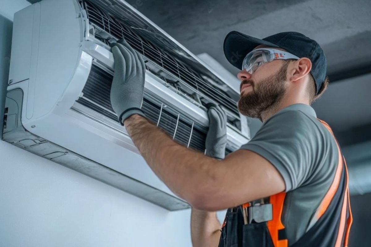 HVAC technician wearing safety glasses and gloves, servicing a wall-mounted air conditioning unit.
