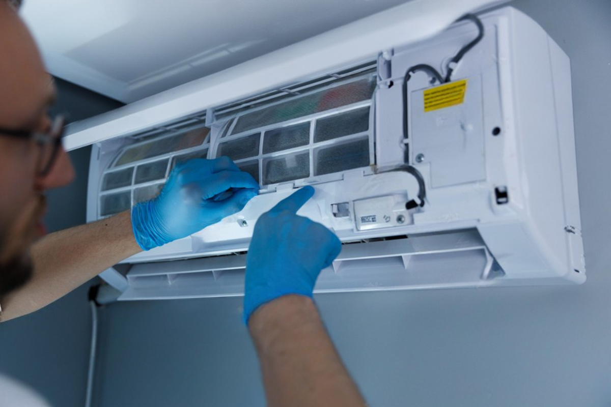 Person in blue gloves cleaning an air conditioner filter. White air conditioner mounted on a gray wall.