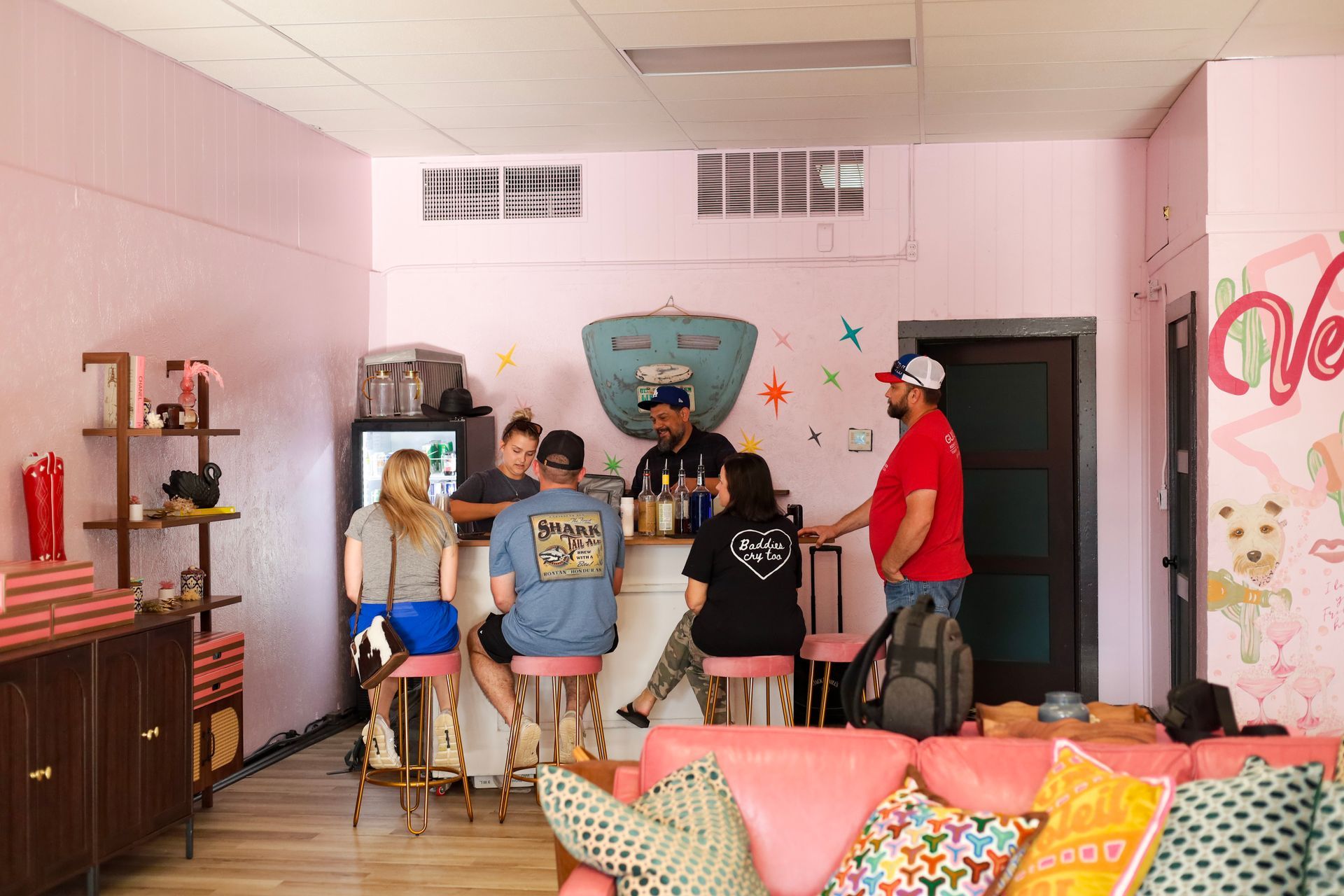 People standing in a pink room around a couch, with shelves, wall art, and bags near the door.