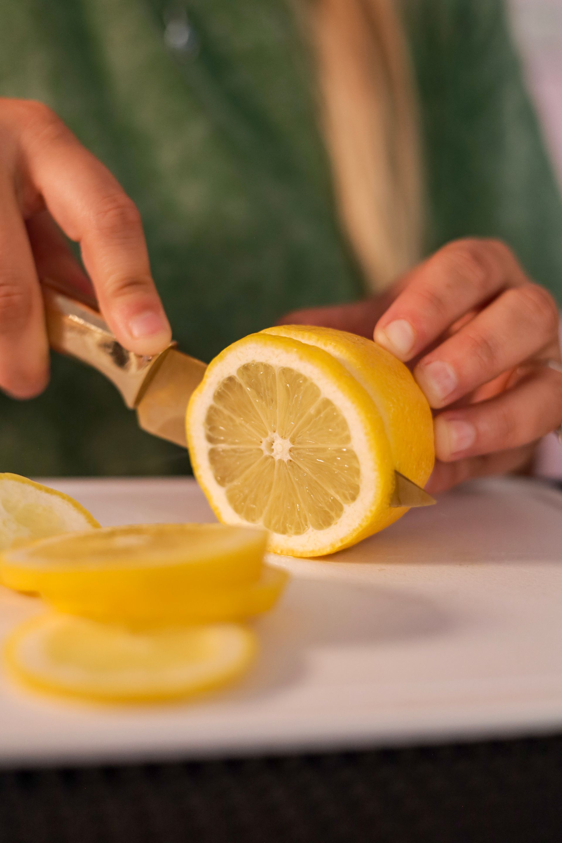 Hands slicing a lemon on a white cutting board, with yellow wedges in the foreground.
