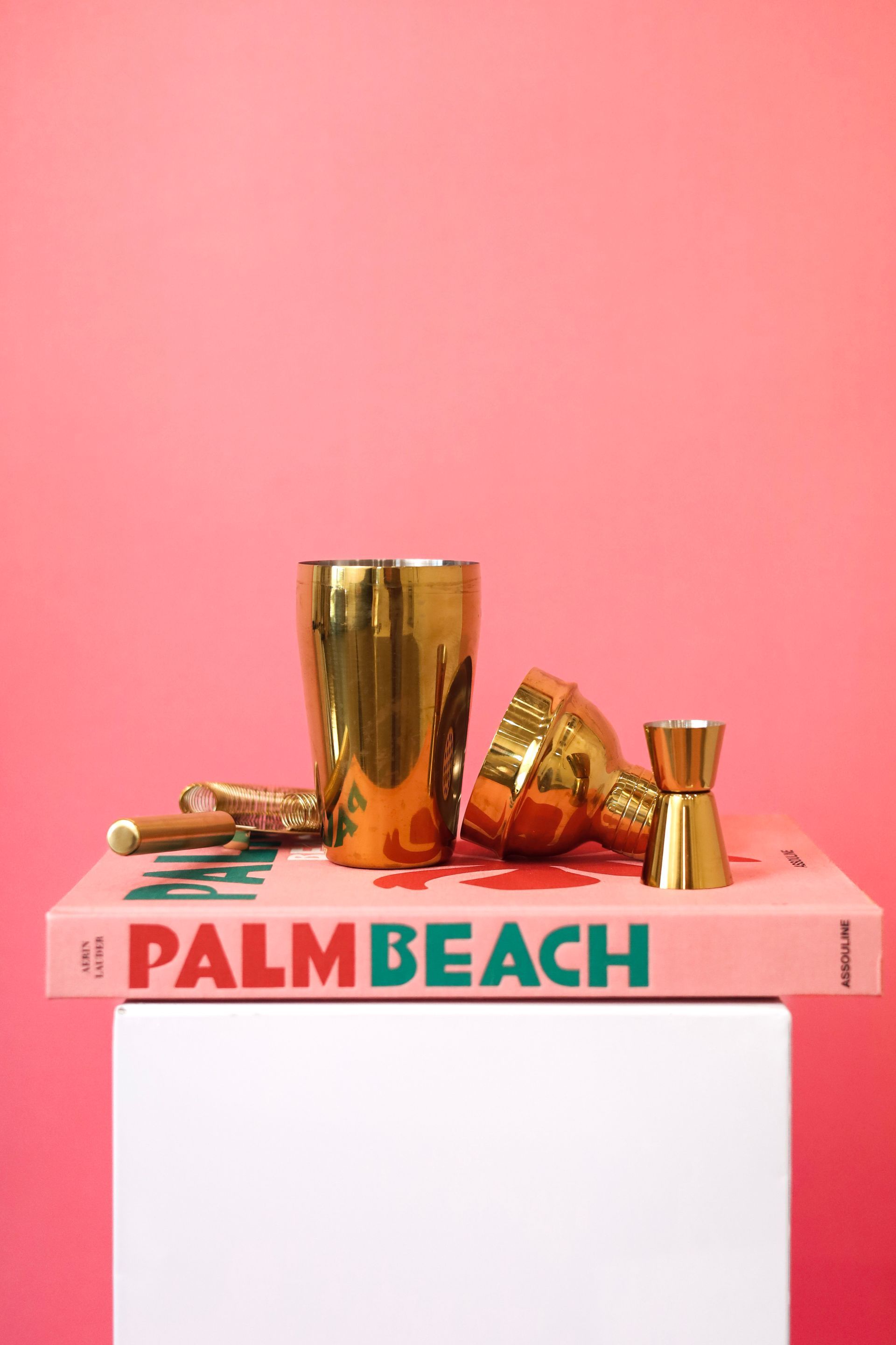 Gold cocktail shaker and glasses on a pink Palm Beach box against a pink background