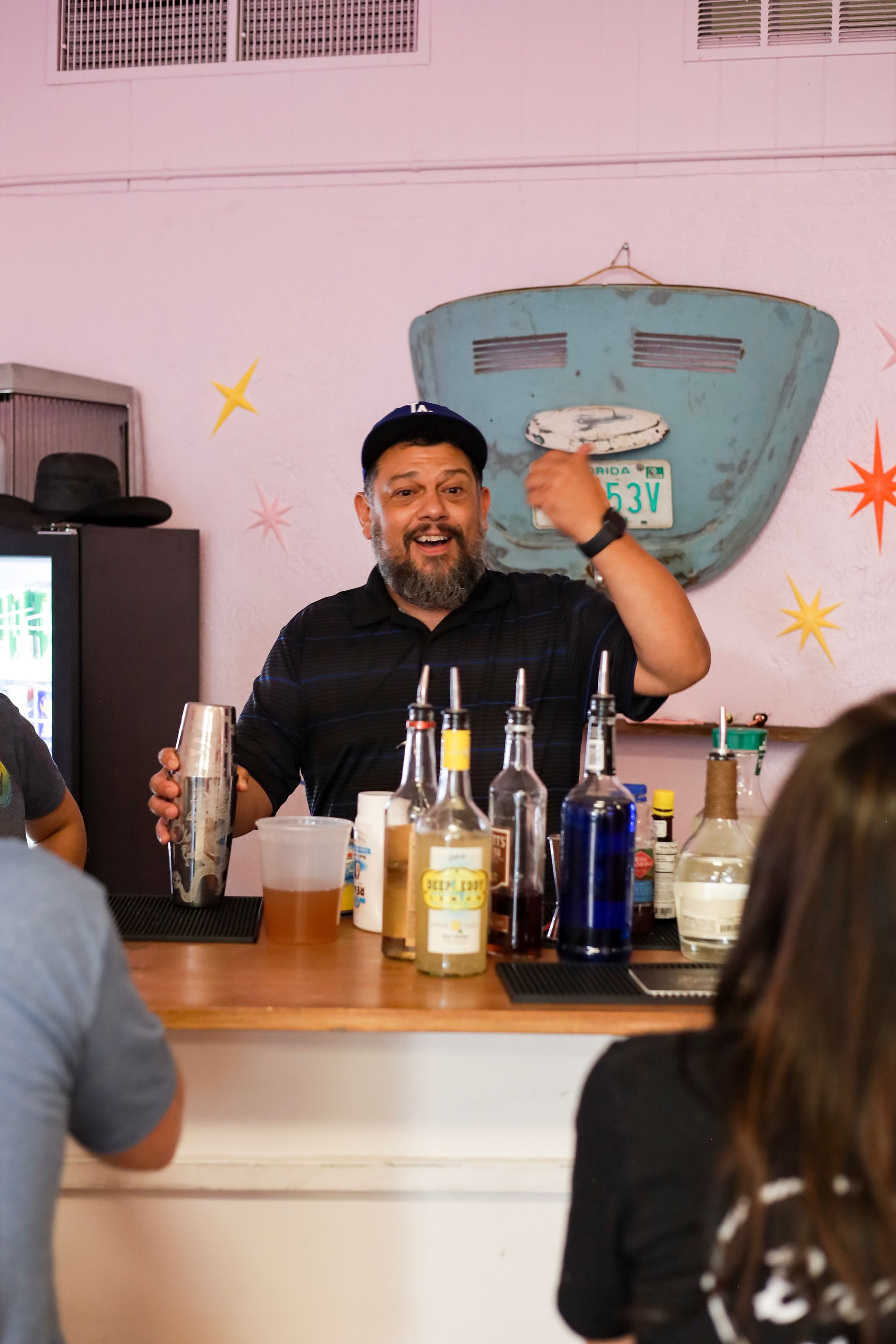 Bartender smiling and raising a drink behind a bar with bottles and colorful wall art.
