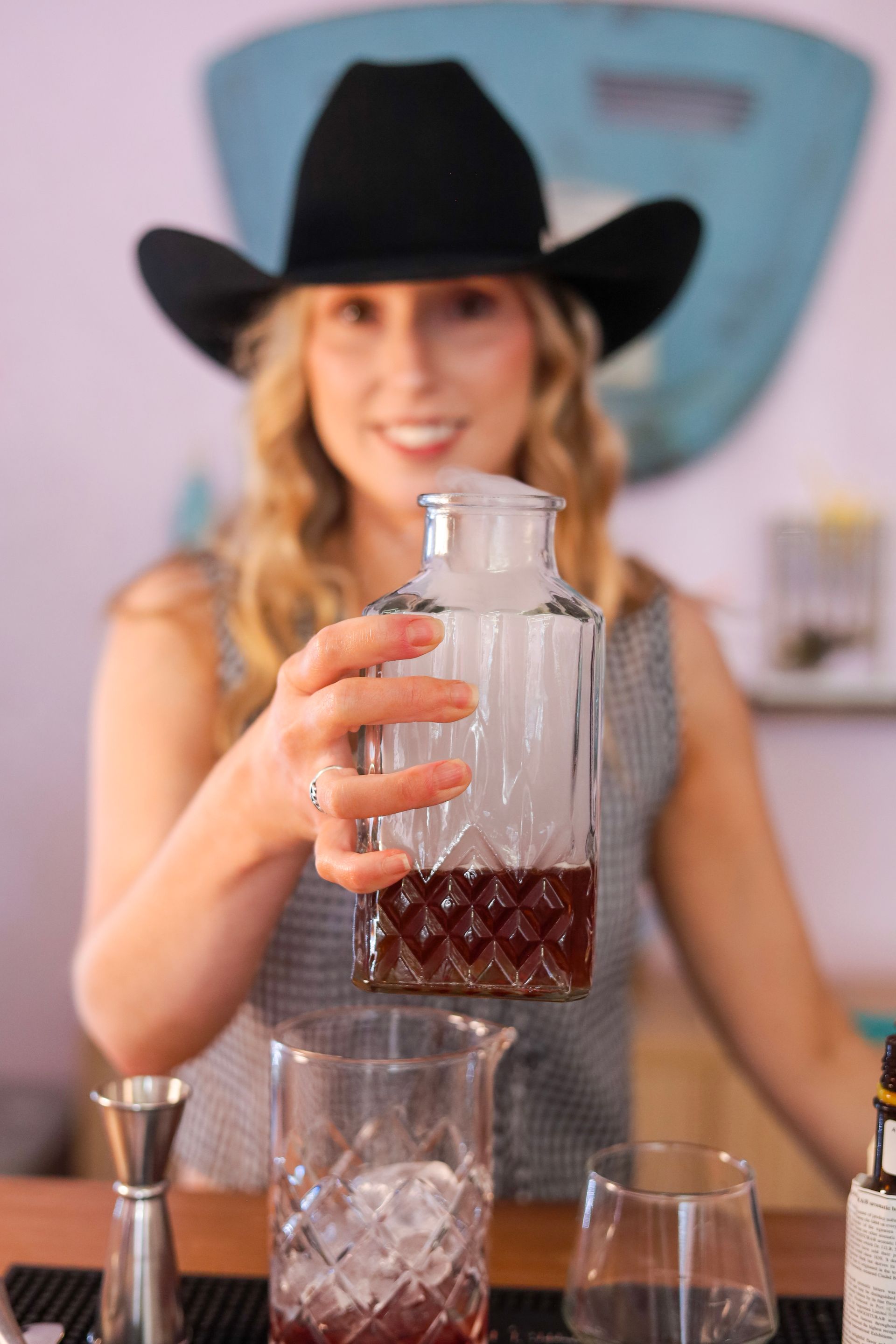 Woman in a black cowboy hat holds a glass cocktail shaker behind a bar.