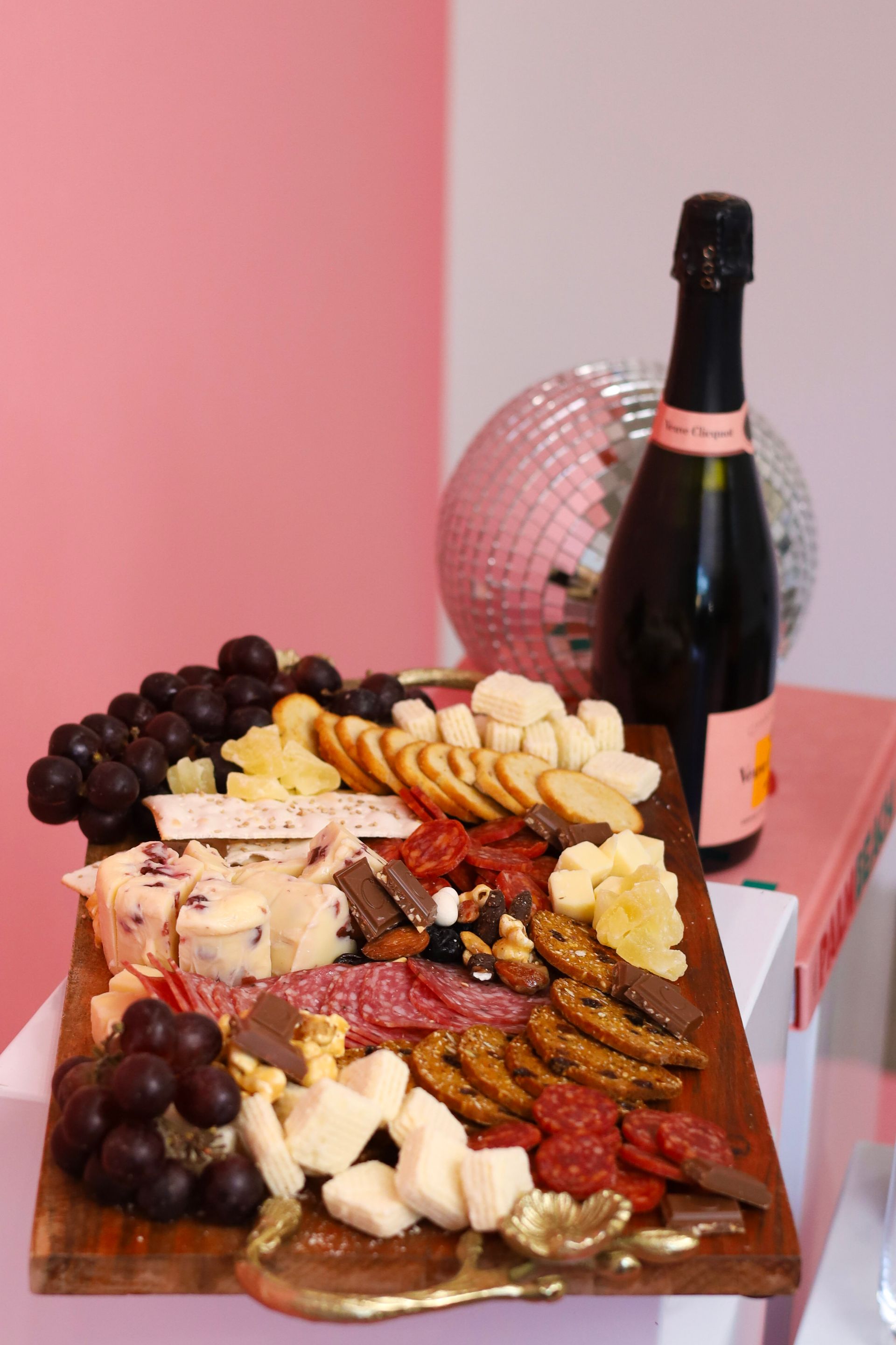 Charcuterie board with grapes, cheese, crackers, nuts, and a bottle of sparkling wine on a pink table