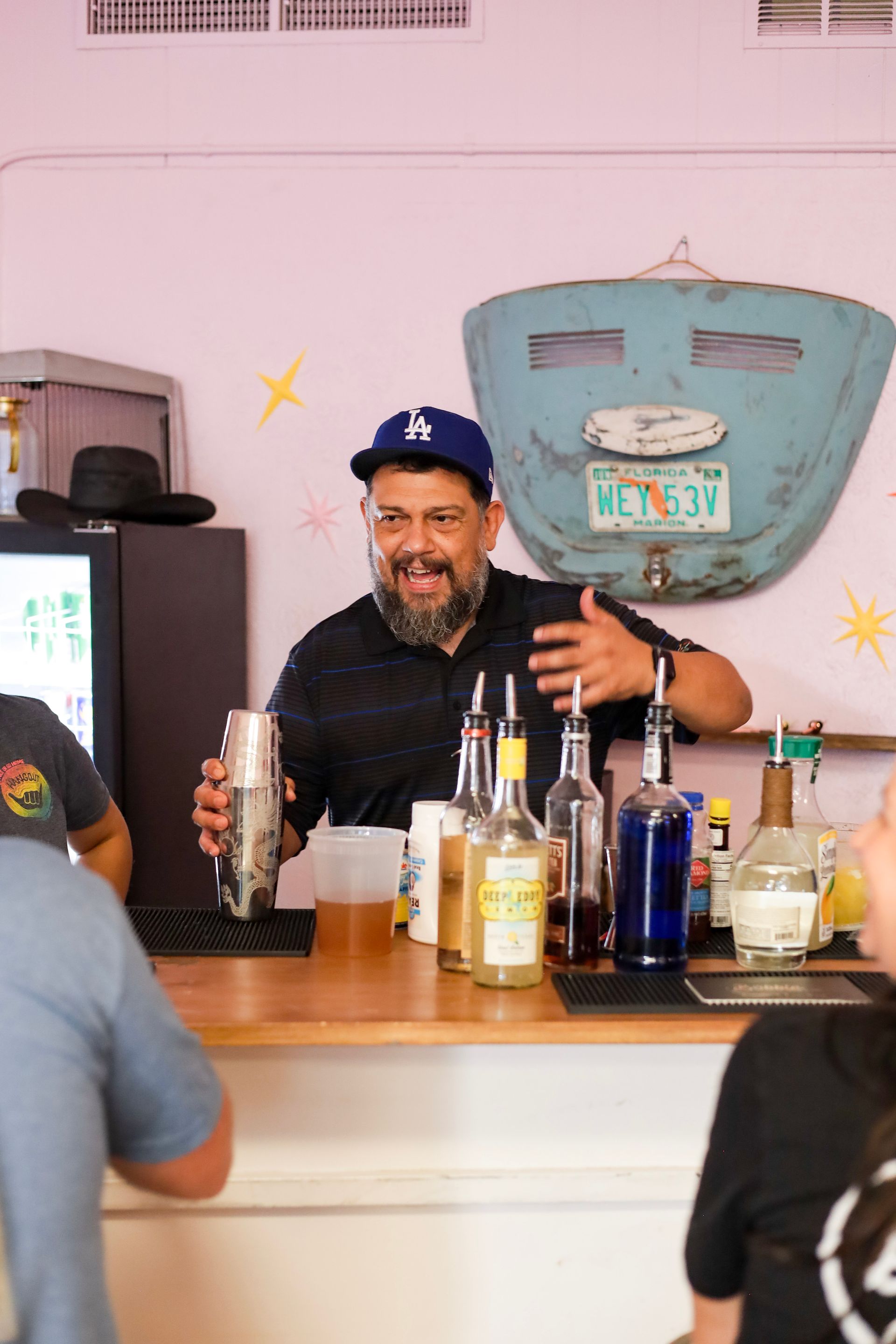 Bartender smiling behind a bar, holding a bottle with assorted liquor bottles displayed in front.