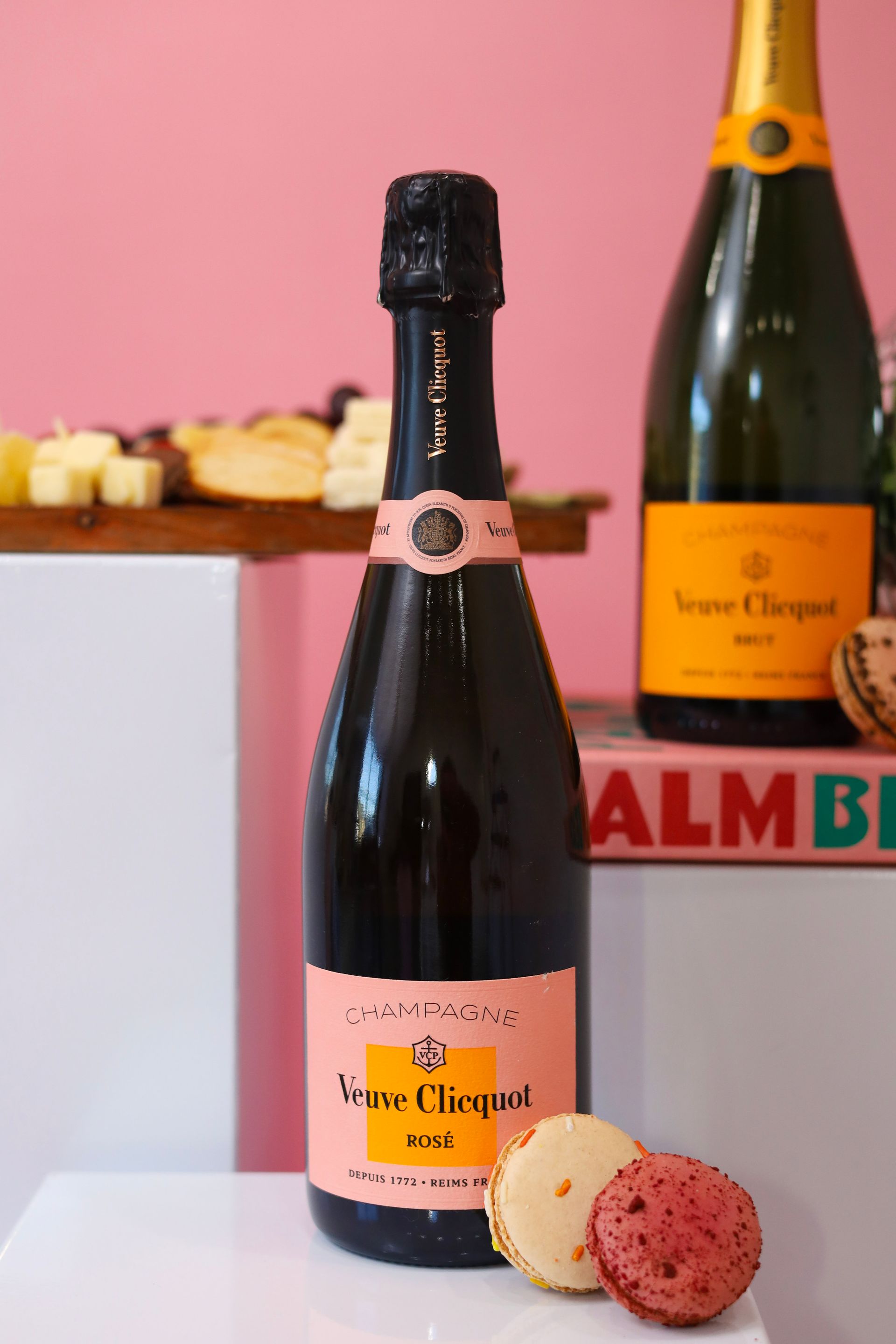 Two champagne bottles with peach labels on a pink table, with macarons in the foreground.