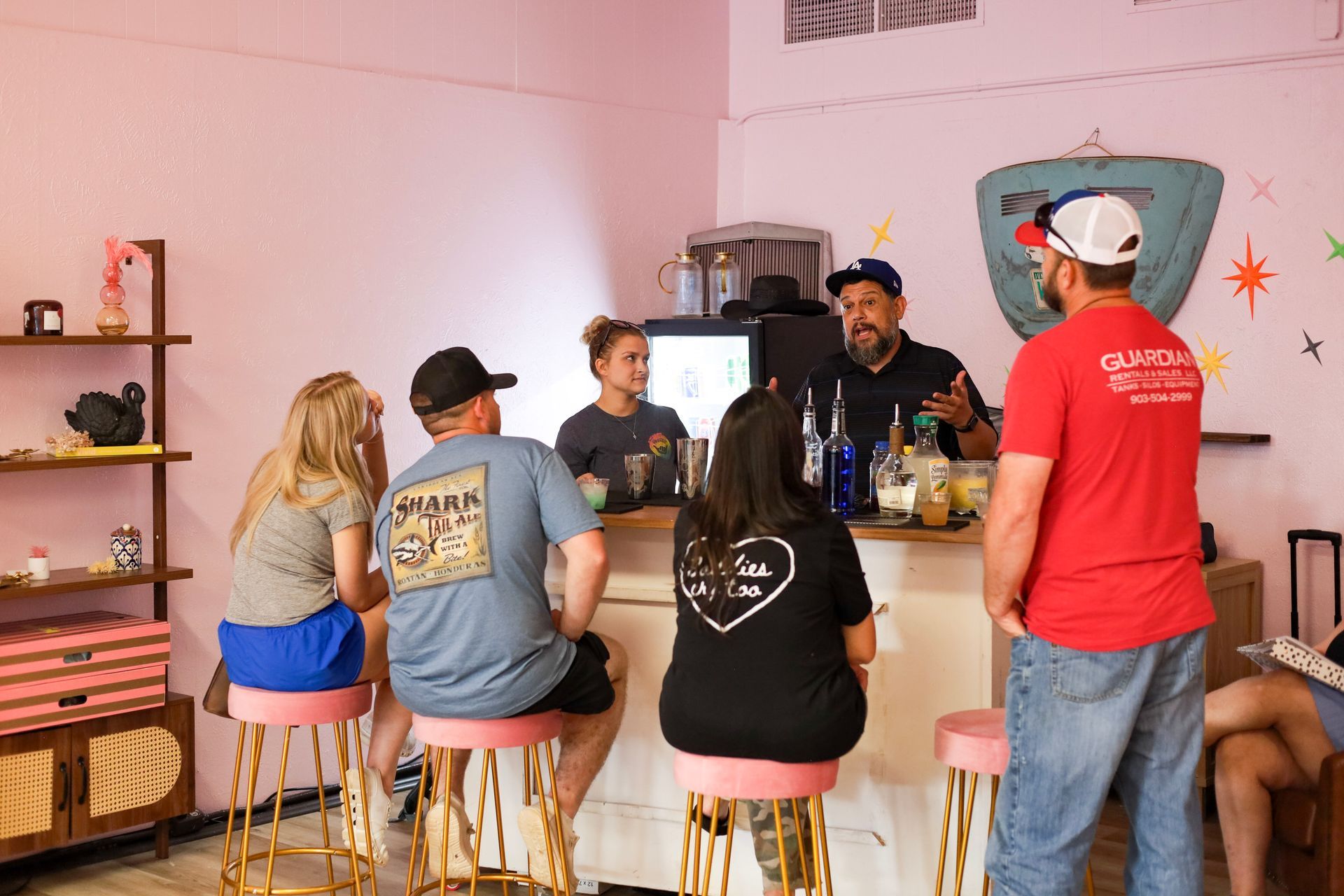 Five people sit at a bar in a pink room, talking with drinks and a bartender behind the counter.