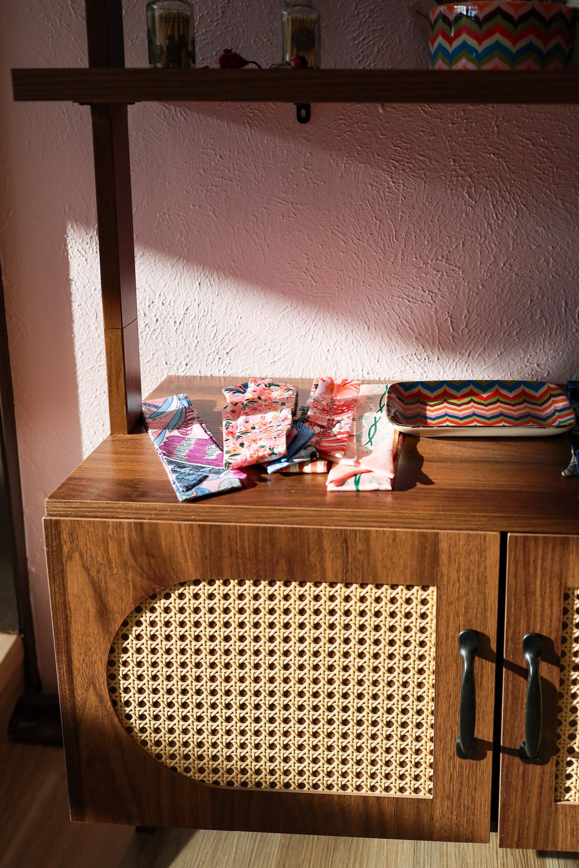 Wooden cabinet with woven panel, holding patterned fabric items on a shelf against a pink wall.