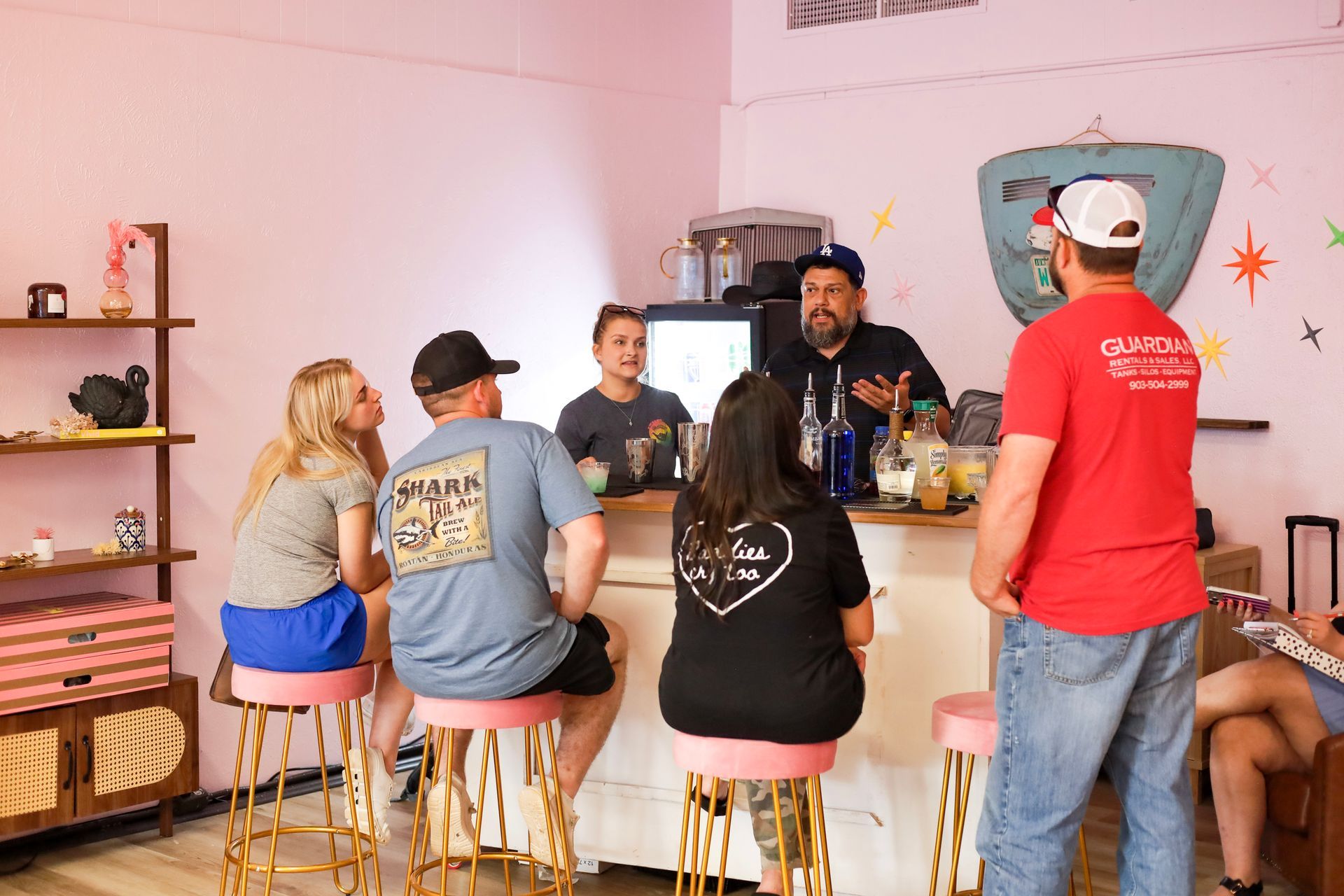 People seated on stools in a casual indoor gathering, listening to a speaker near a decorated wall.