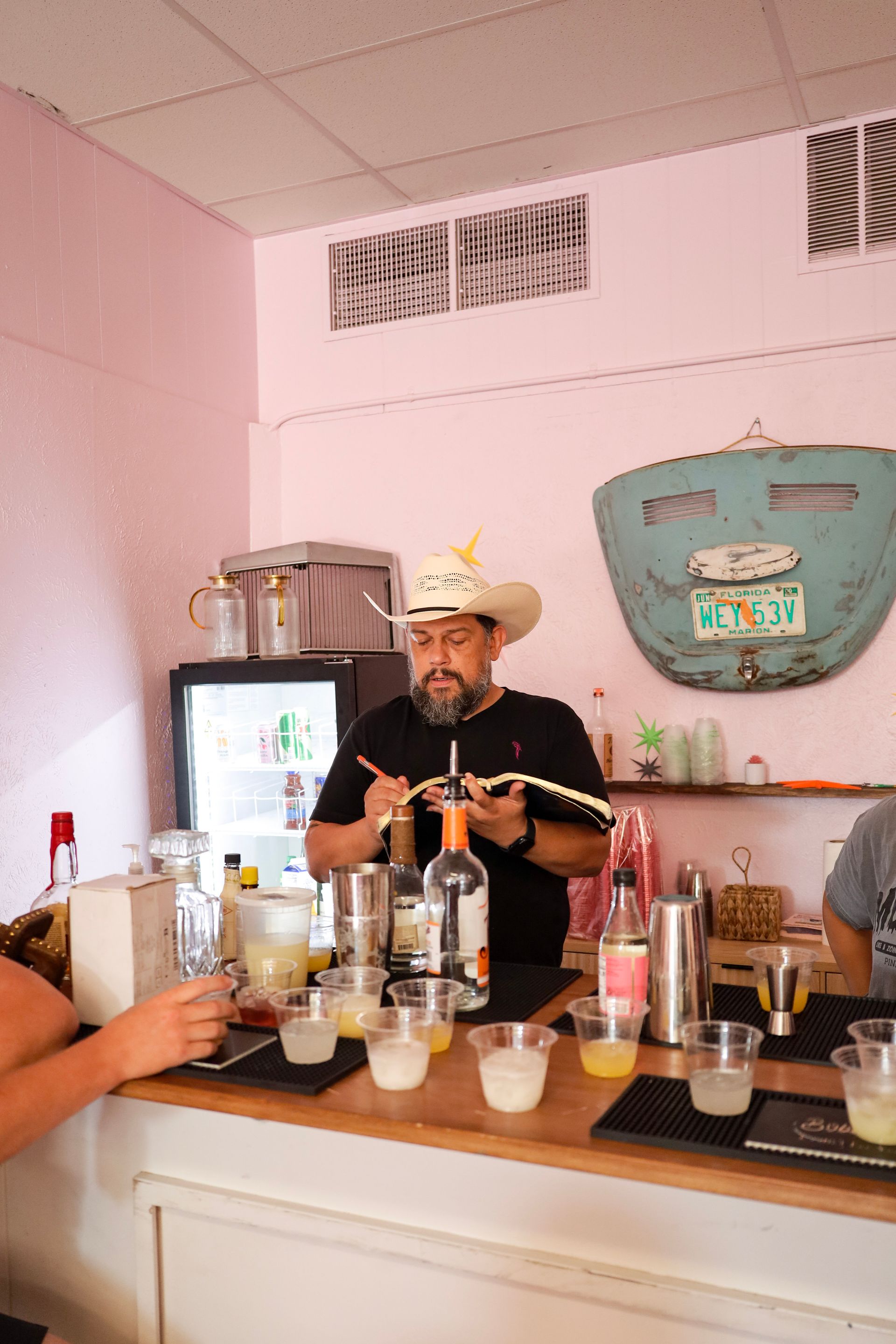 Bartender mixing drinks behind a pink bar with a large green cooler and several bottles