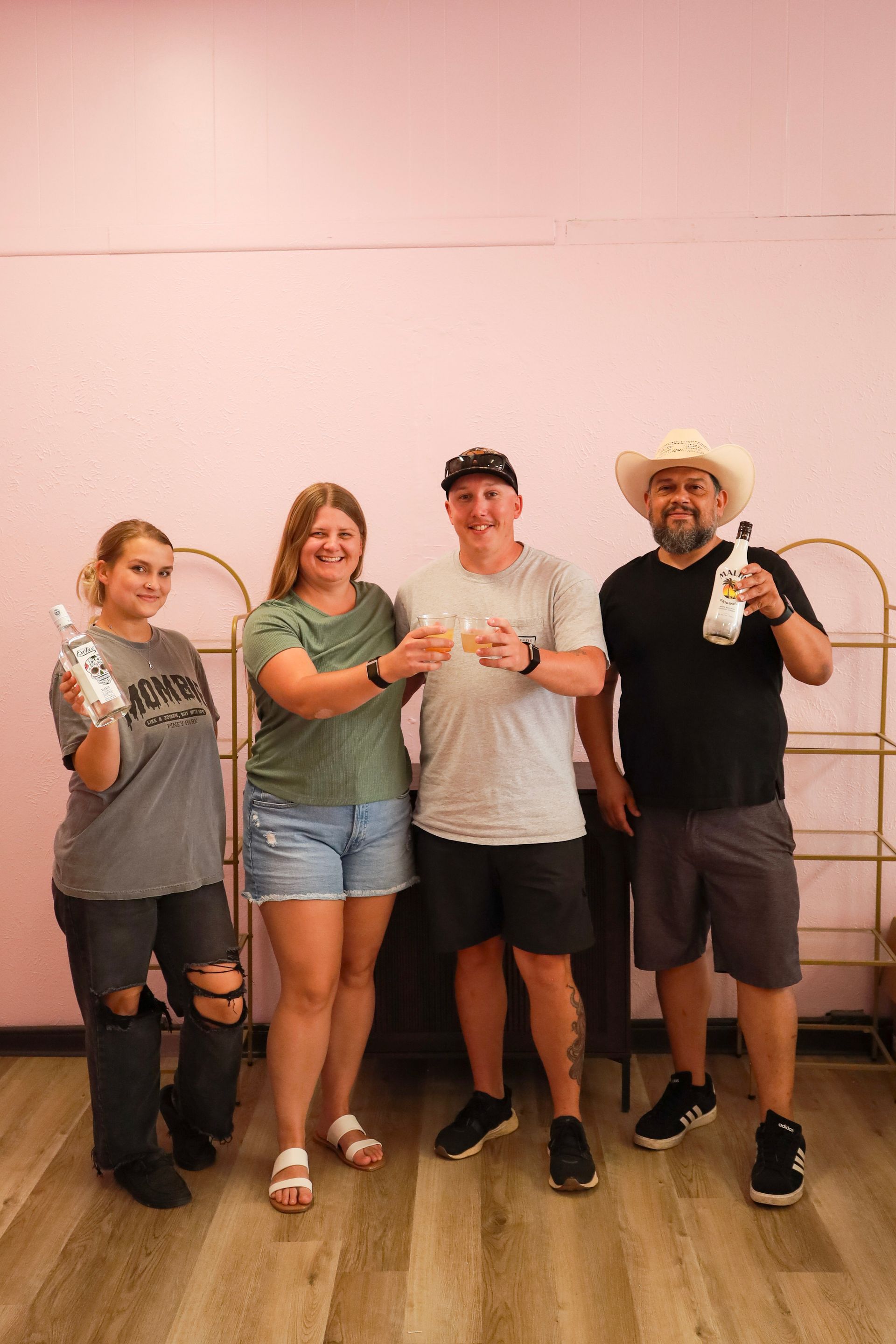 Four people posing indoors with drinks, standing in front of a pink wall and wicker chairs.