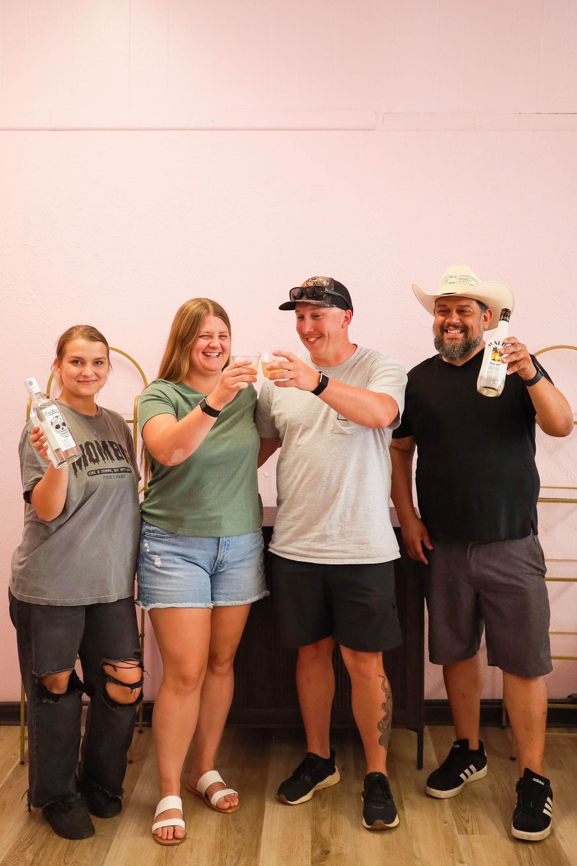 Four people posing indoors, smiling and holding drinks, two wearing hats, in casual clothes