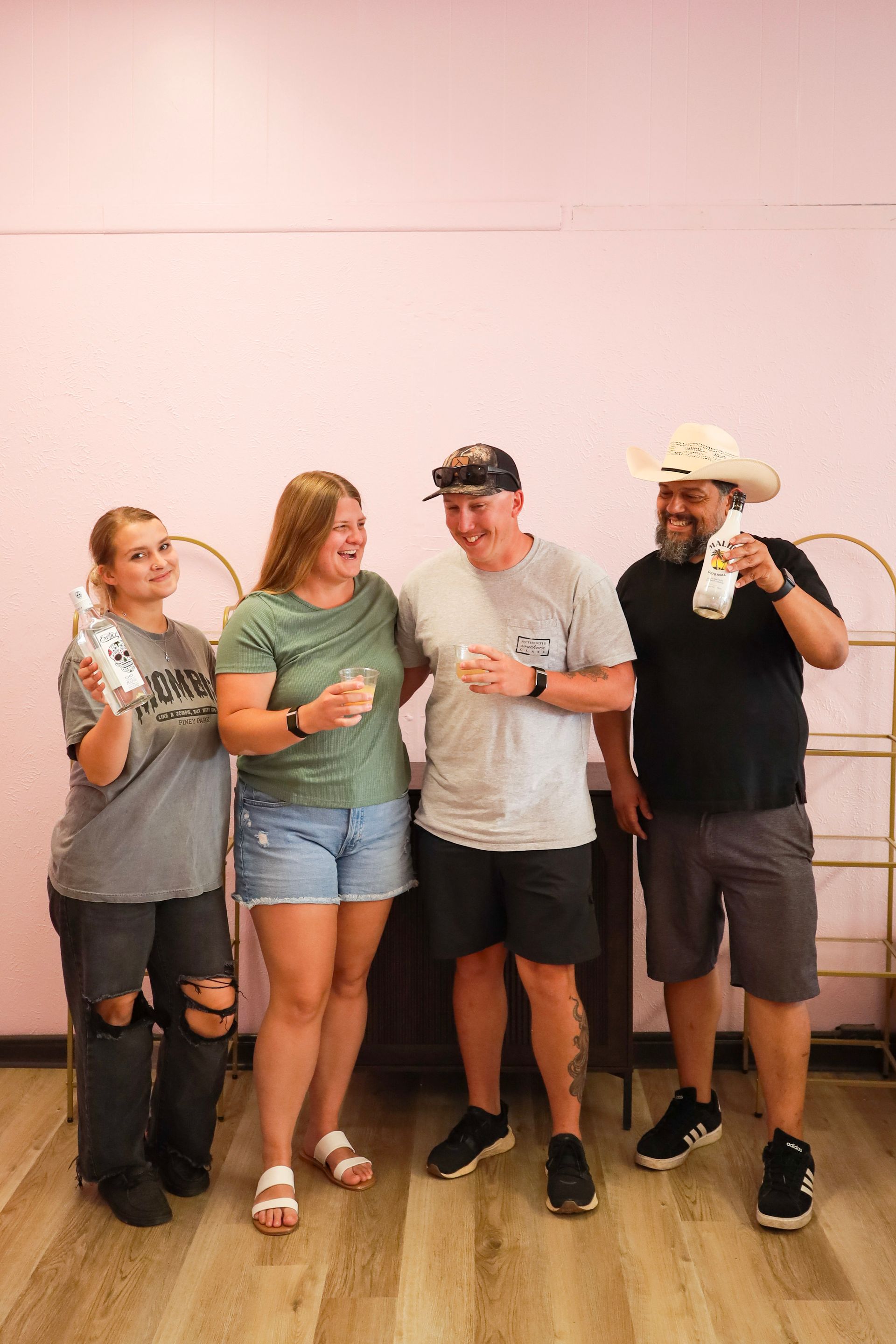 Four people standing indoors against a pale wall, holding drinks and smiling at the camera.