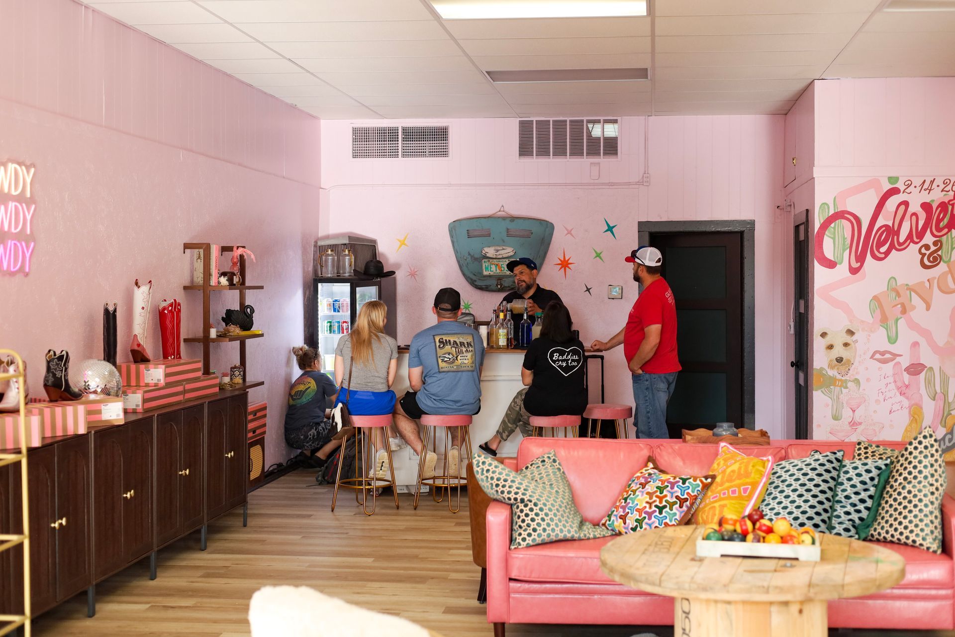People gathered in a pink lounge with a bar, sofas, and wall art, chatting and standing near a counter.