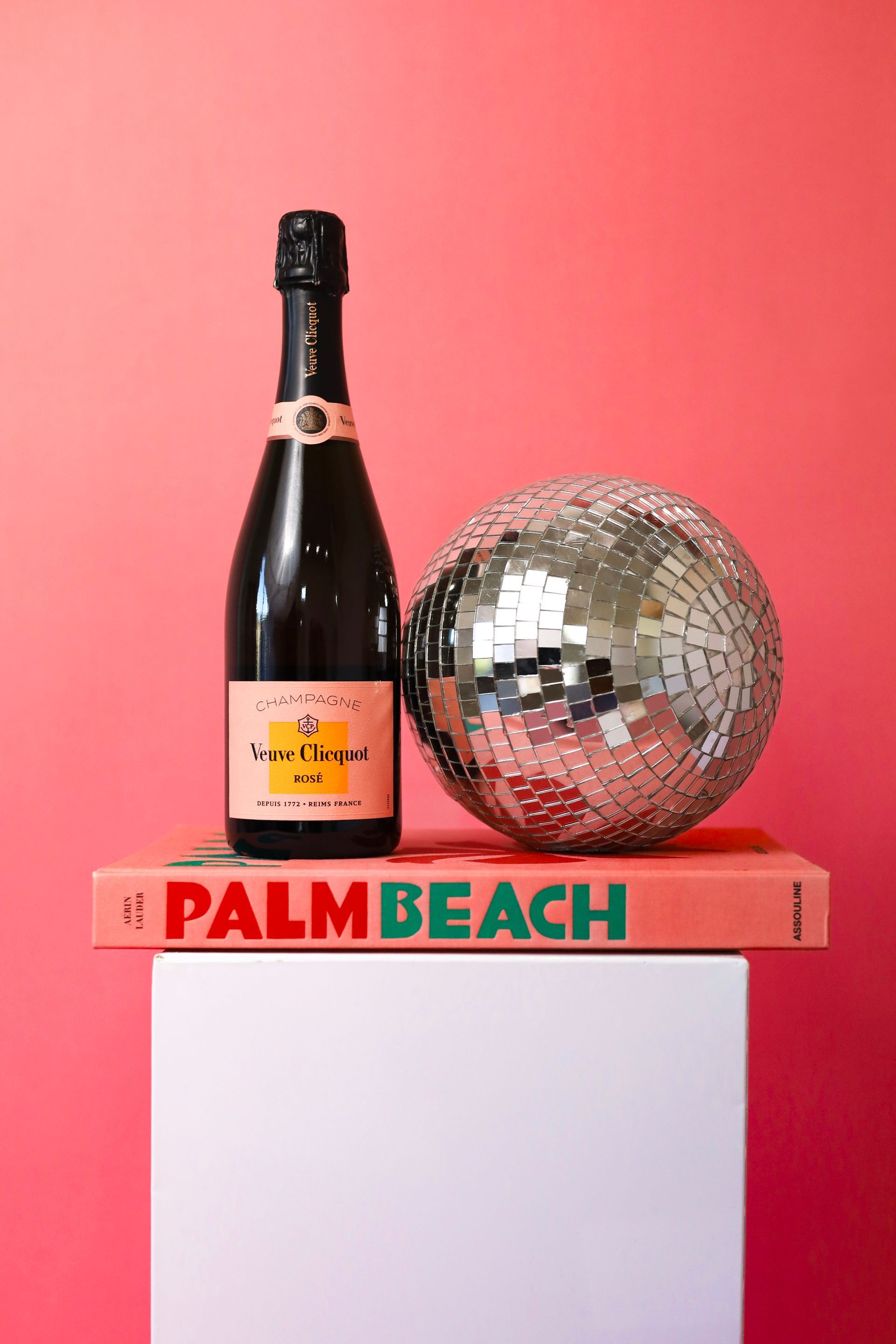 Champagne bottle and silver disco ball on a pink Palm Beach book against a coral background