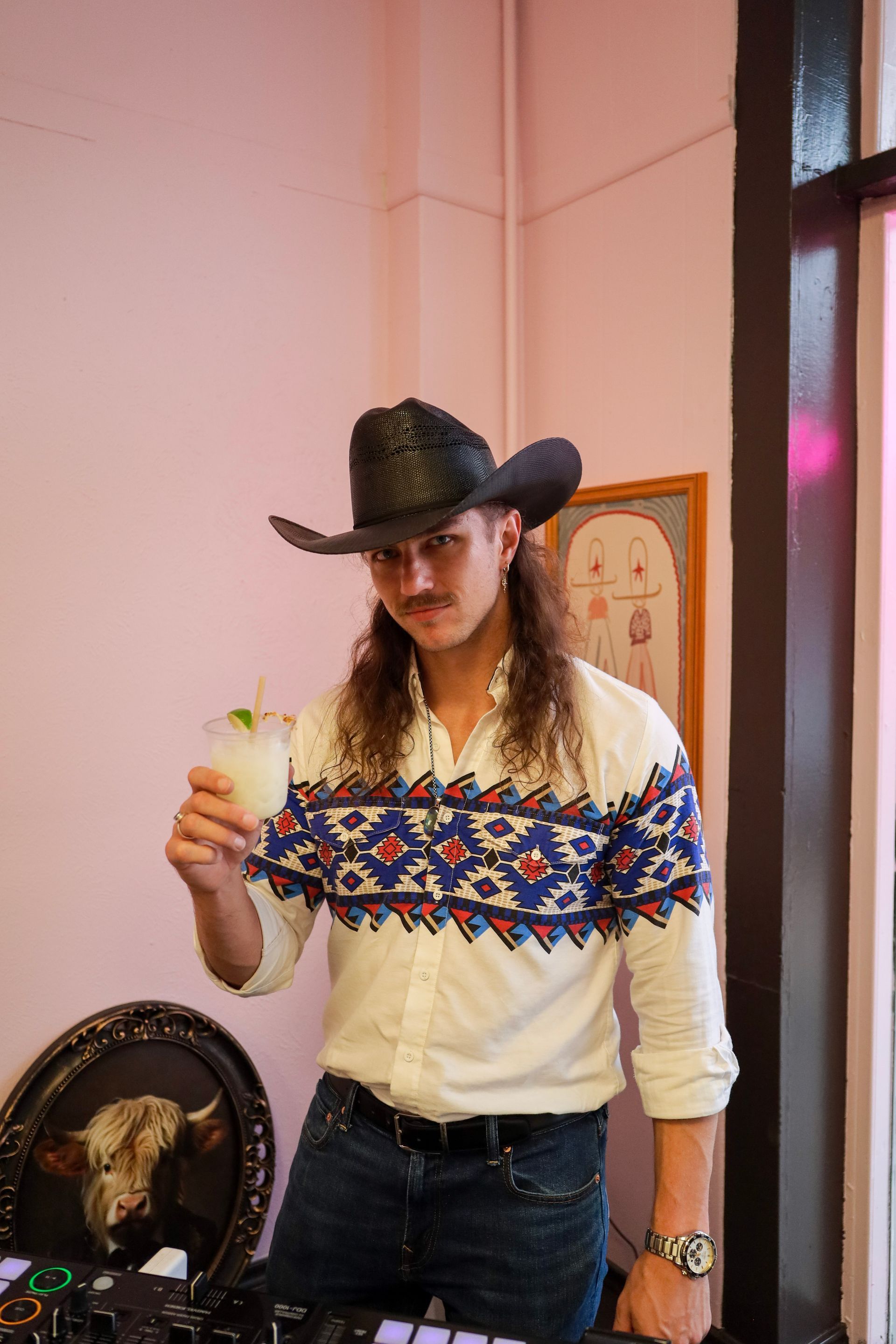 Person in a black cowboy hat holding a drink indoors beside a window and decorative wall art
