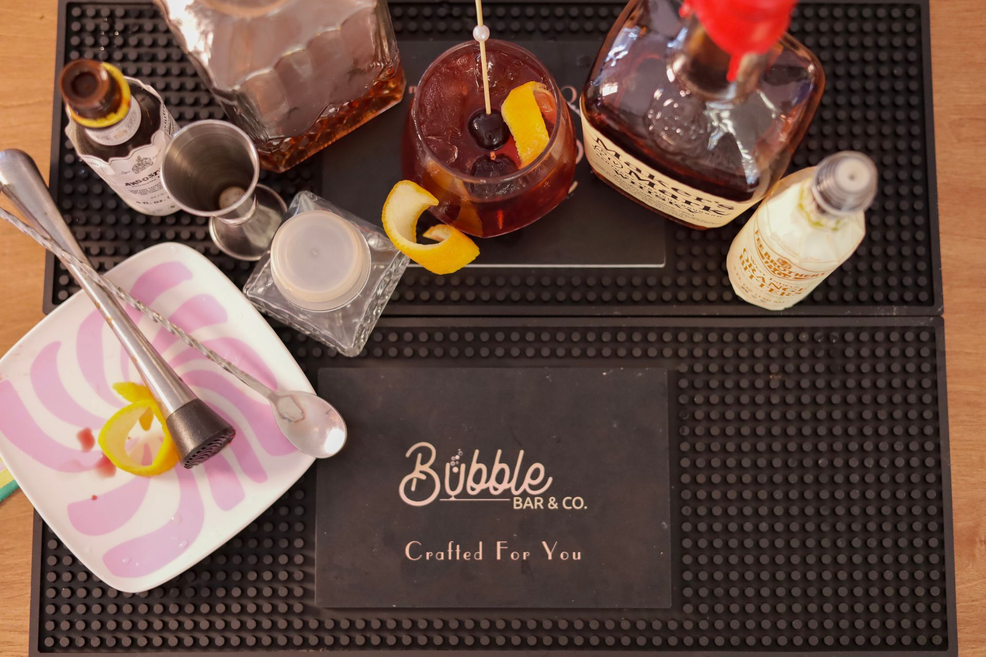 Assorted liquor bottles and bar tools on a black mat labeled “Bubble Club Co. Crafted For You.”