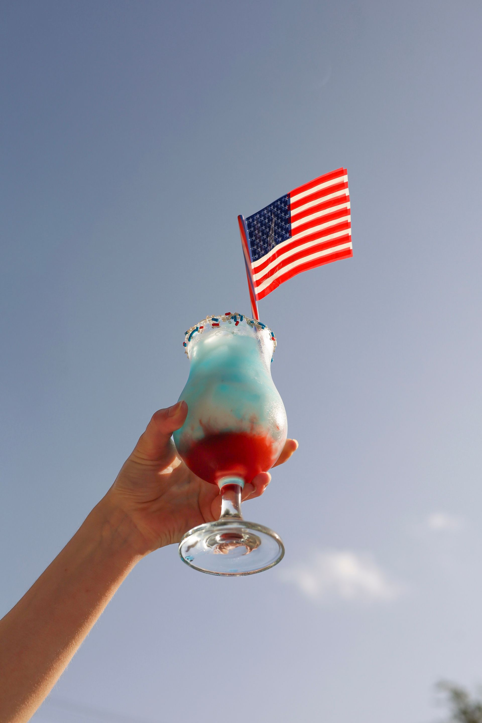Hand holding a red, white, and blue cocktail topped with whipped cream and a small American flag against the sky