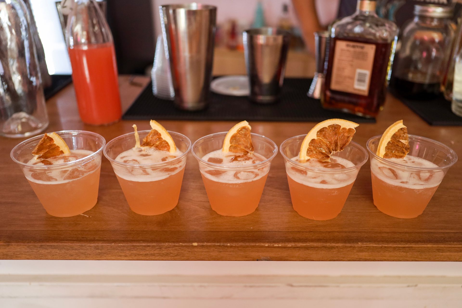 Five orange cocktails lined on a wooden bar with garnishes and bar tools behind them