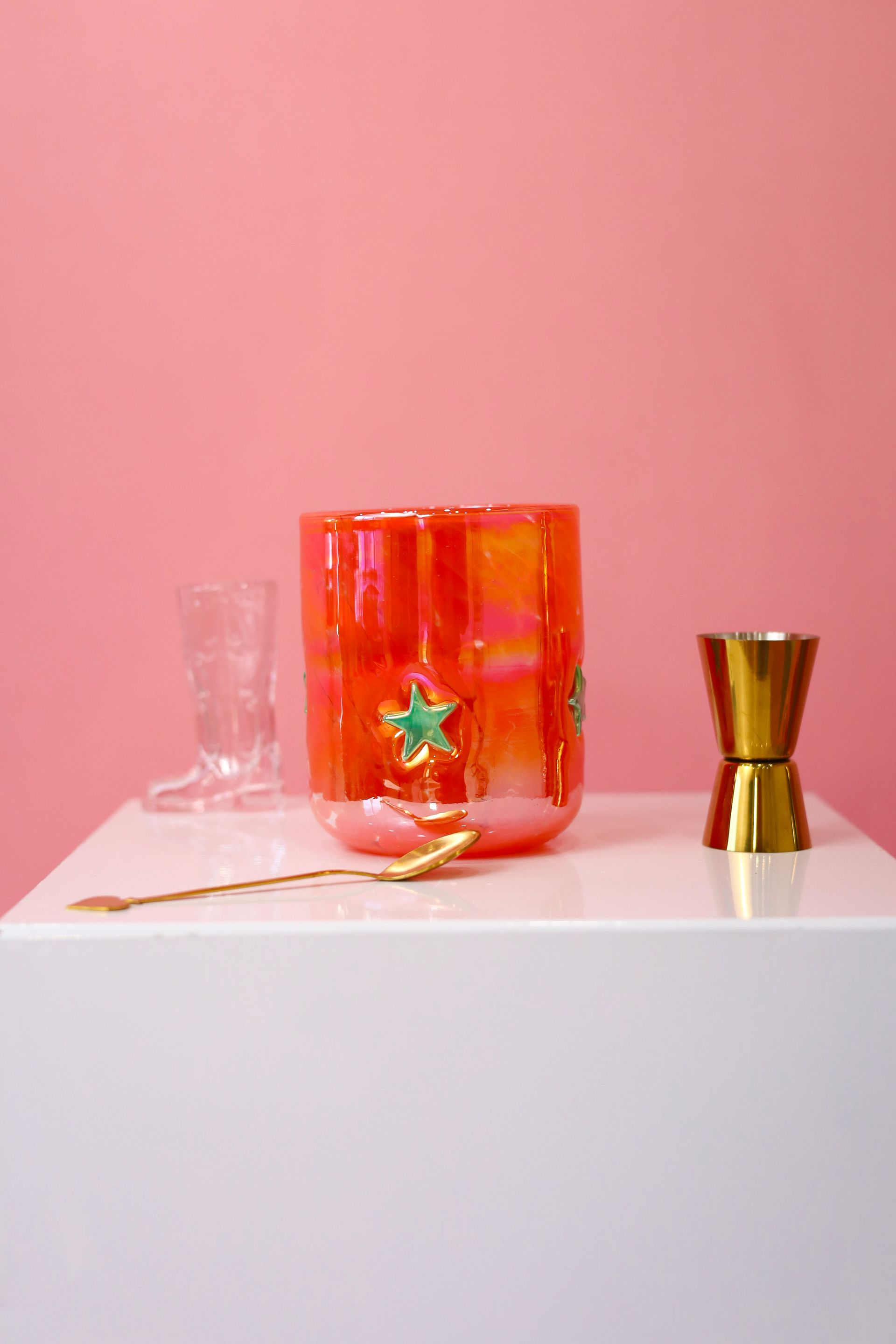 Pink background with a white pedestal holding a red-orange glass, a clear flute, and a gold shot glass