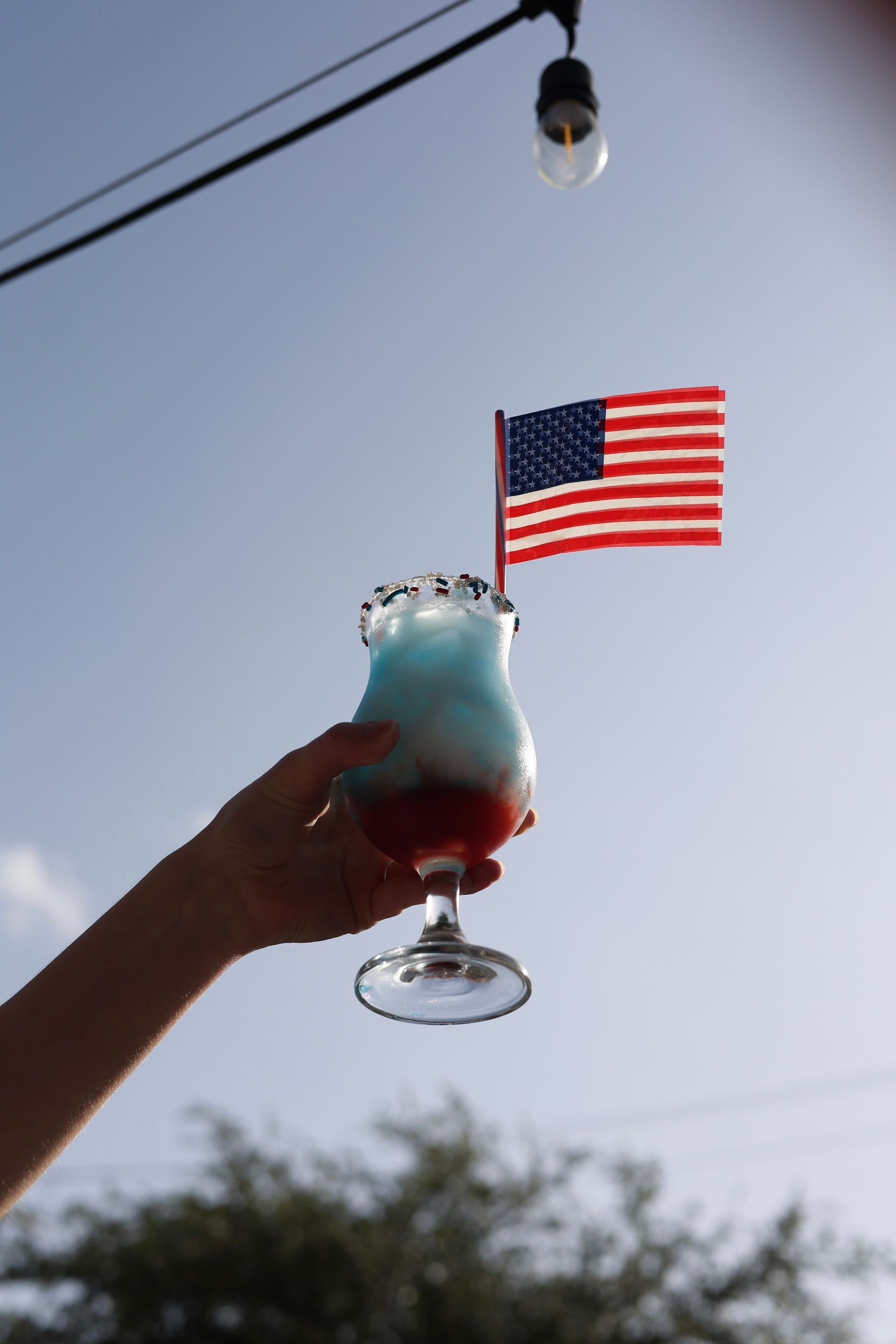 Hand holding a red, white, and blue frozen drink beneath an American flag against a clear sky