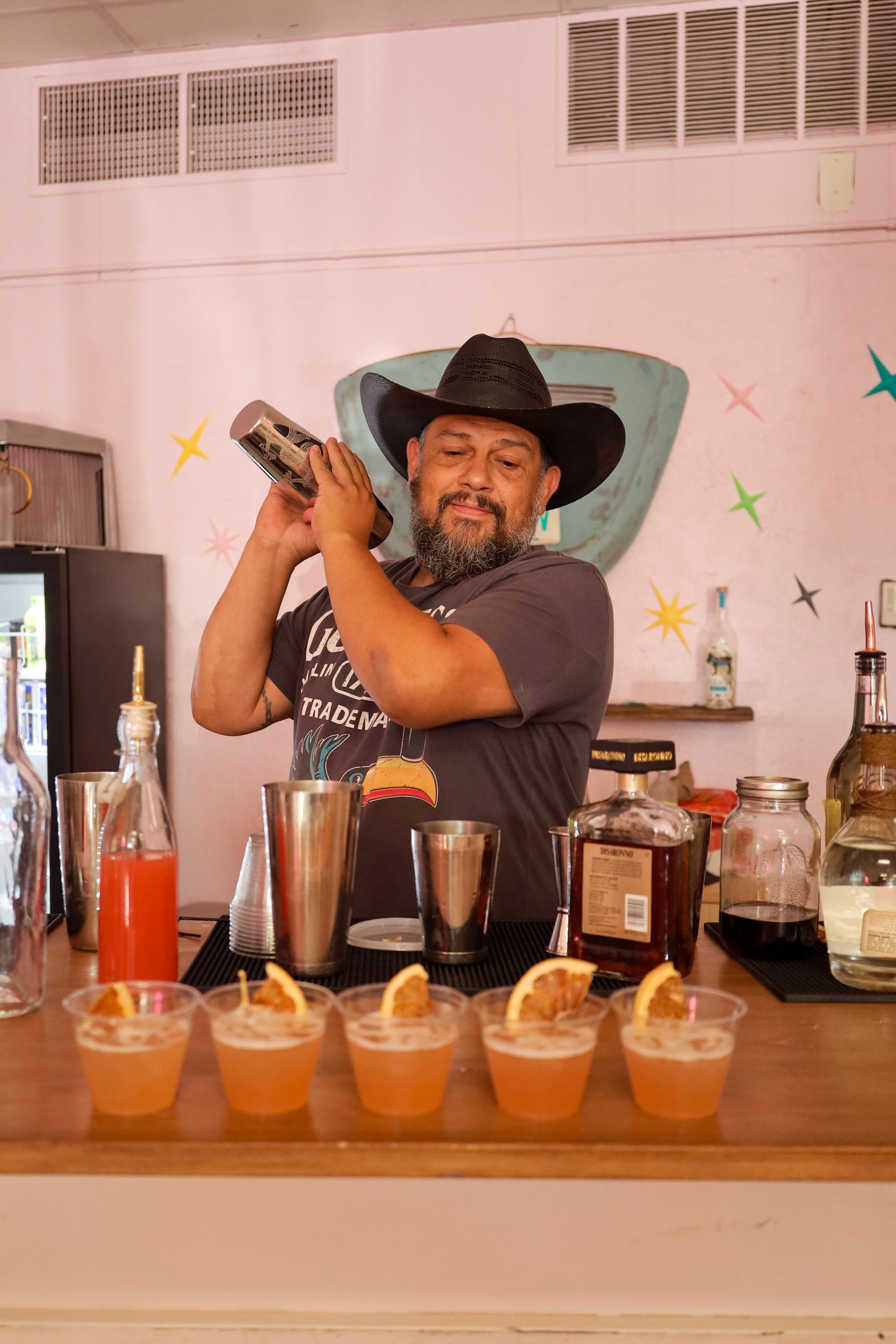 Bartender in a cowboy hat shaking a cocktail behind a bar with several drinks lined up in front.