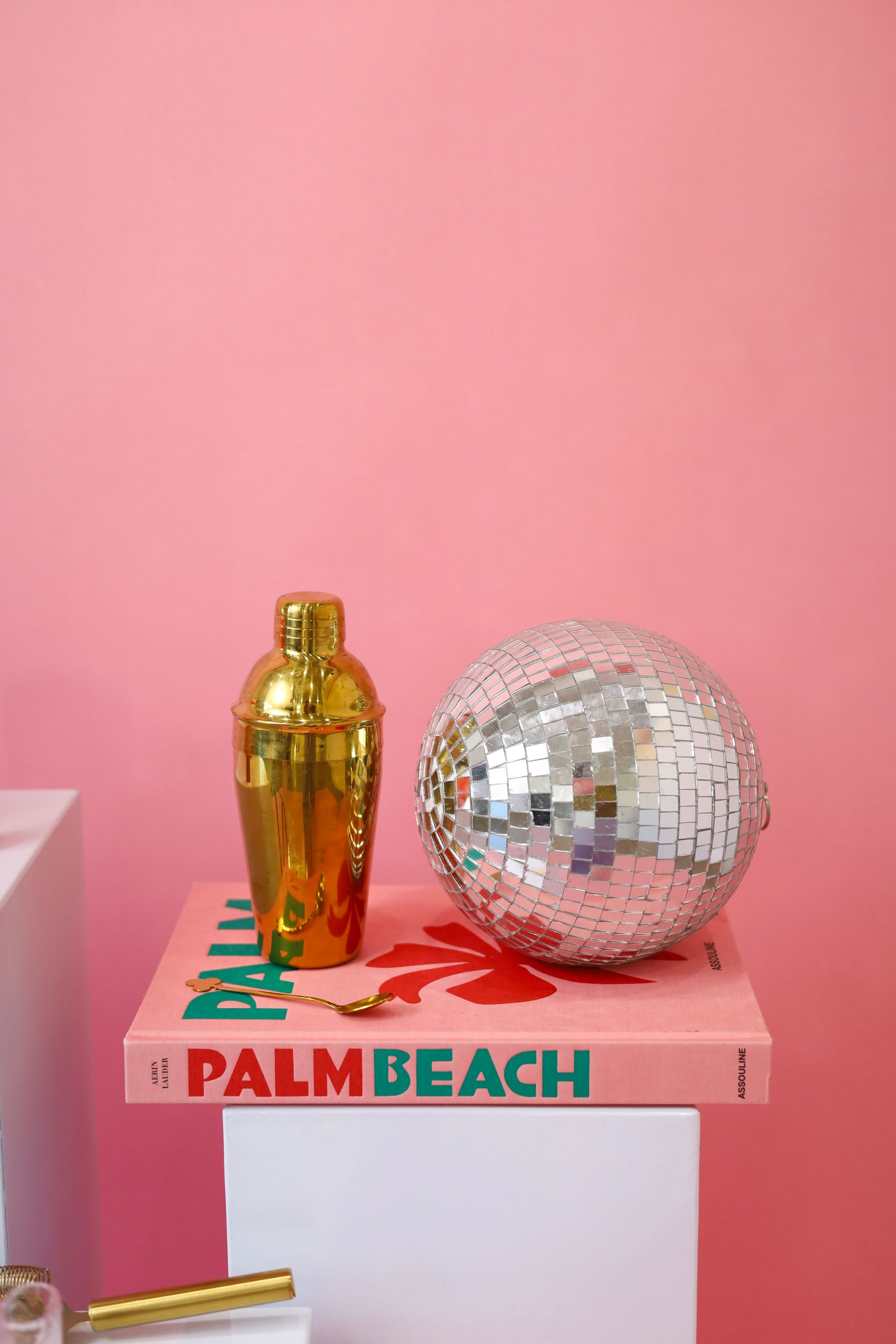 Gold cocktail shaker and disco ball on a pink Palm Beach book against a pink background
