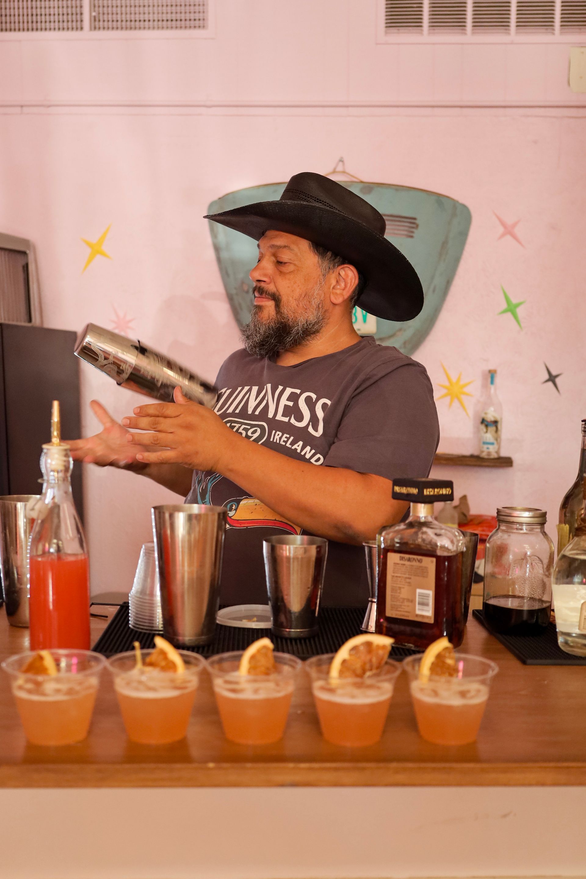 Bartender in black hat shaking a cocktail behind a bar with rows of colorful drinks and bottles