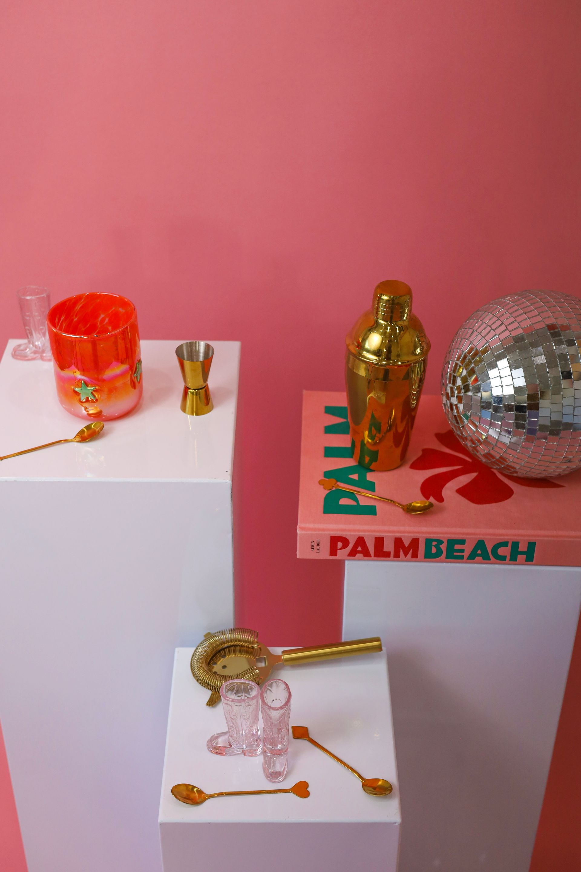 Decorative display of gold and silver bottles and disco balls on white pedestals against a pink wall