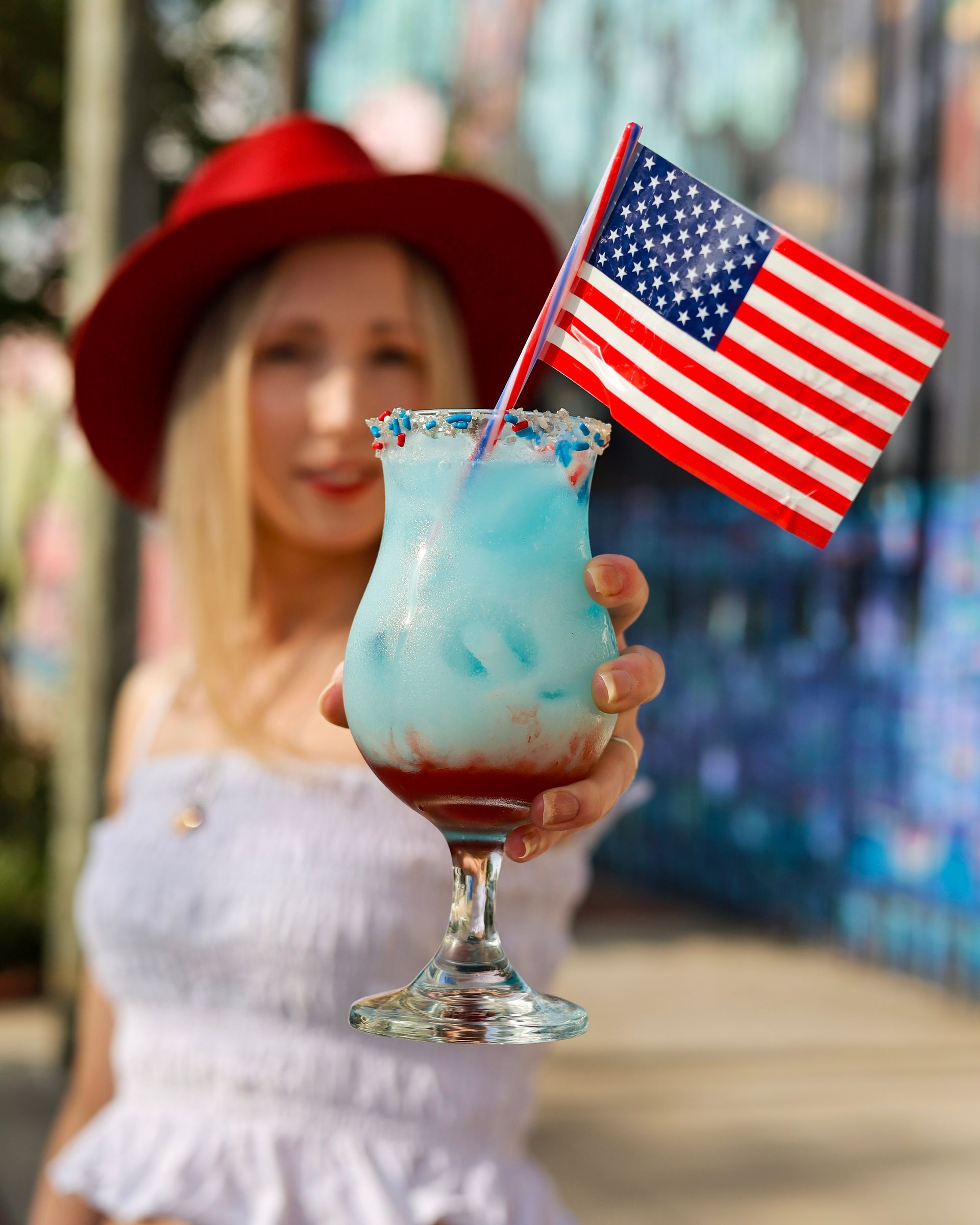 Woman in red hat holding a blue-red patriotic cocktail topped with an American flag outdoors