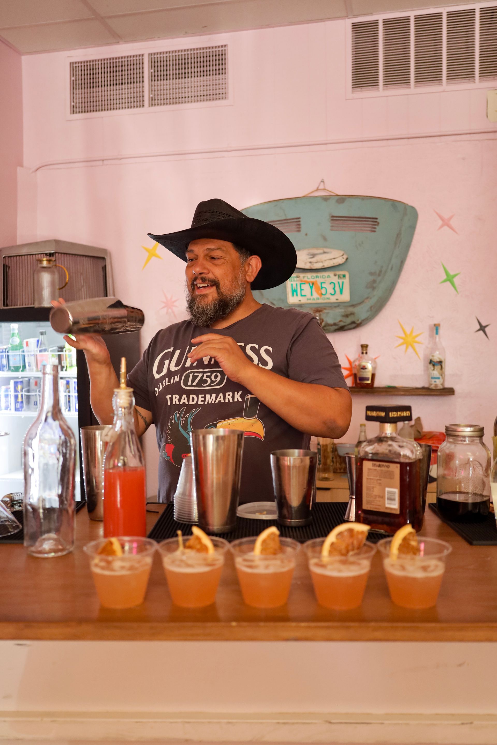 Bartender in black hat behind a bar, pouring drinks with colorful cocktails lined up in front