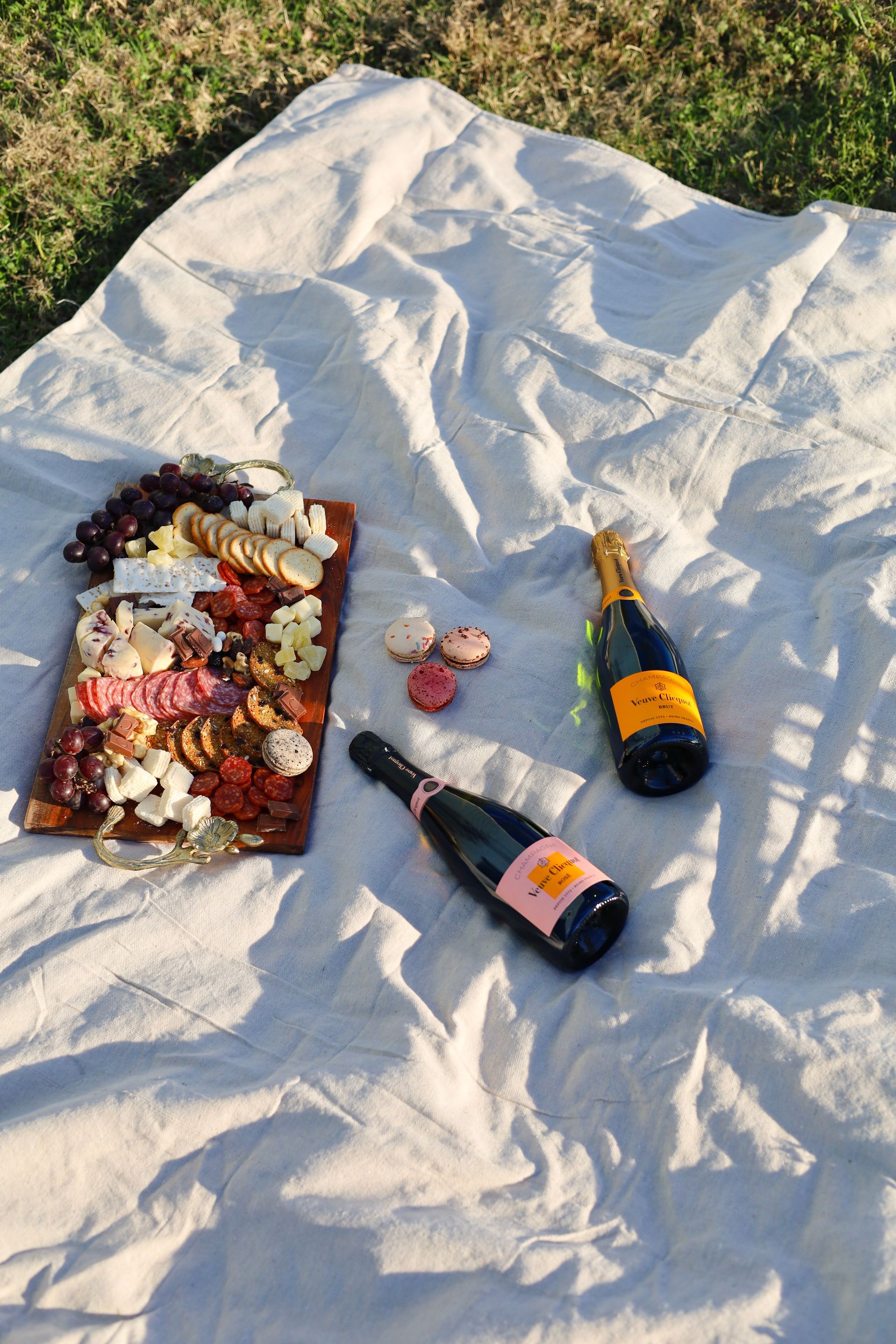 Picnic blanket with wine bottles, food, and a charcuterie board on grass.