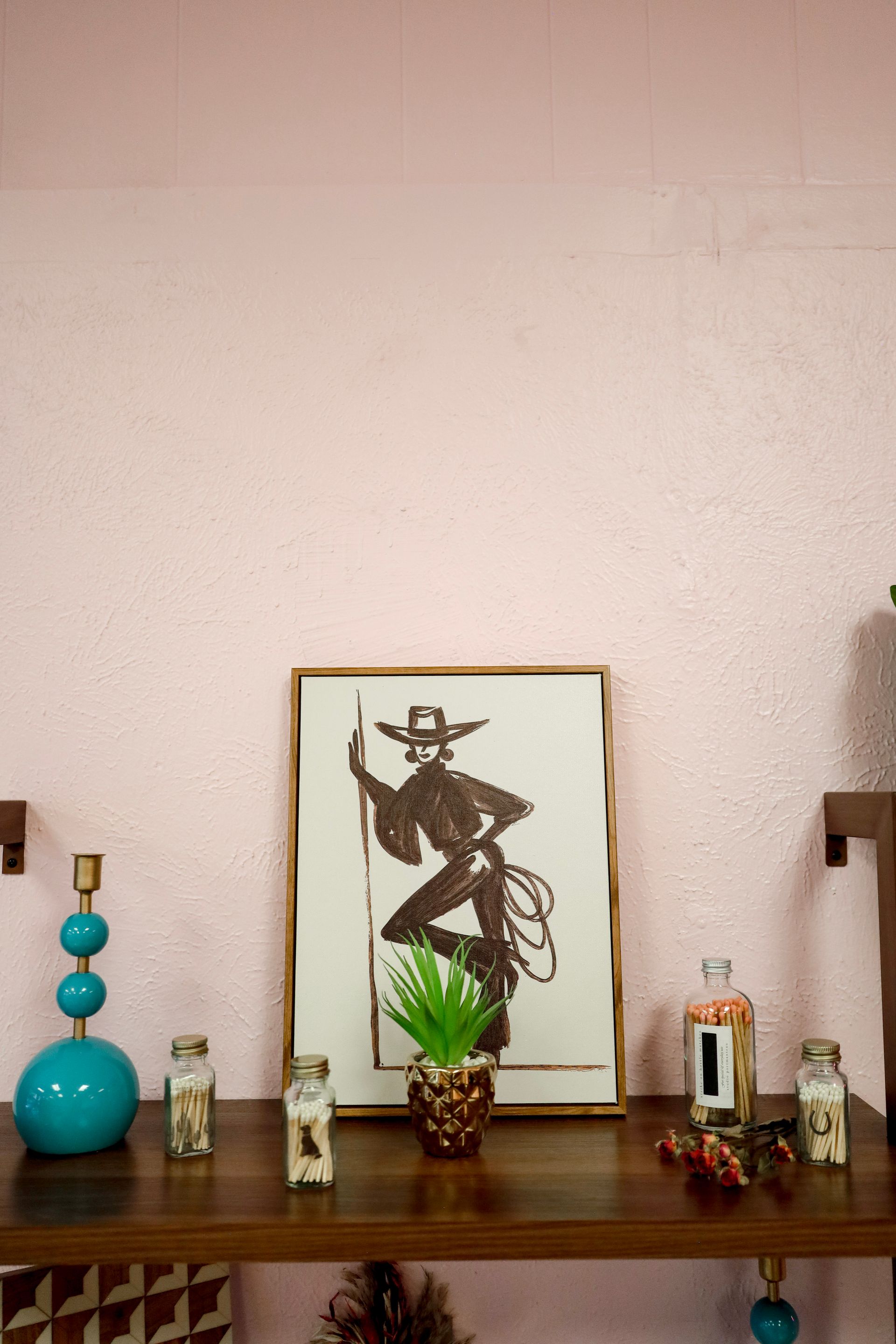 Decorative shelf with framed artwork, teal vase, small plants, and candles against a pale wall