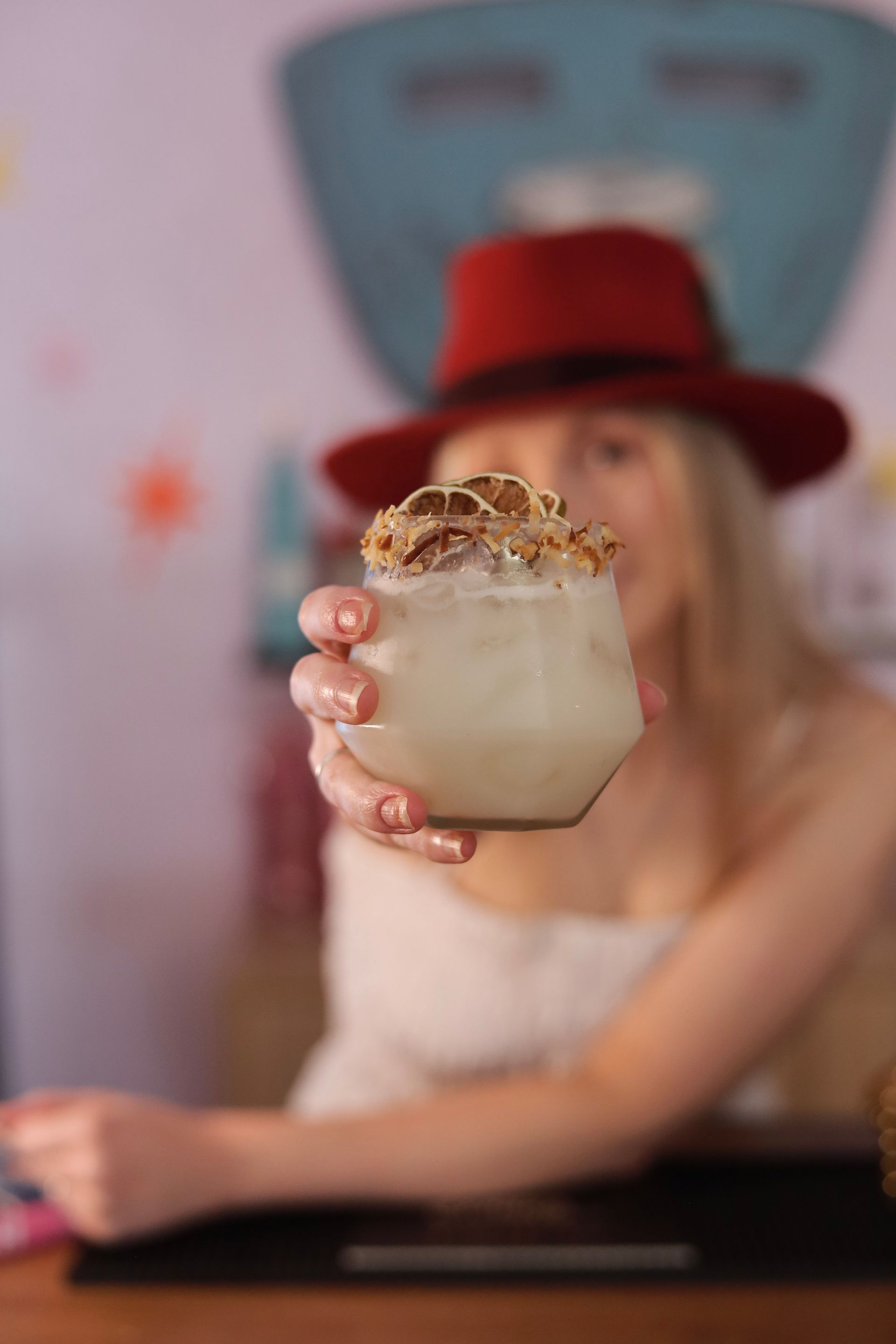 A hand holding a cocktail with ice and garnish in front of a blurred person in a red hat.