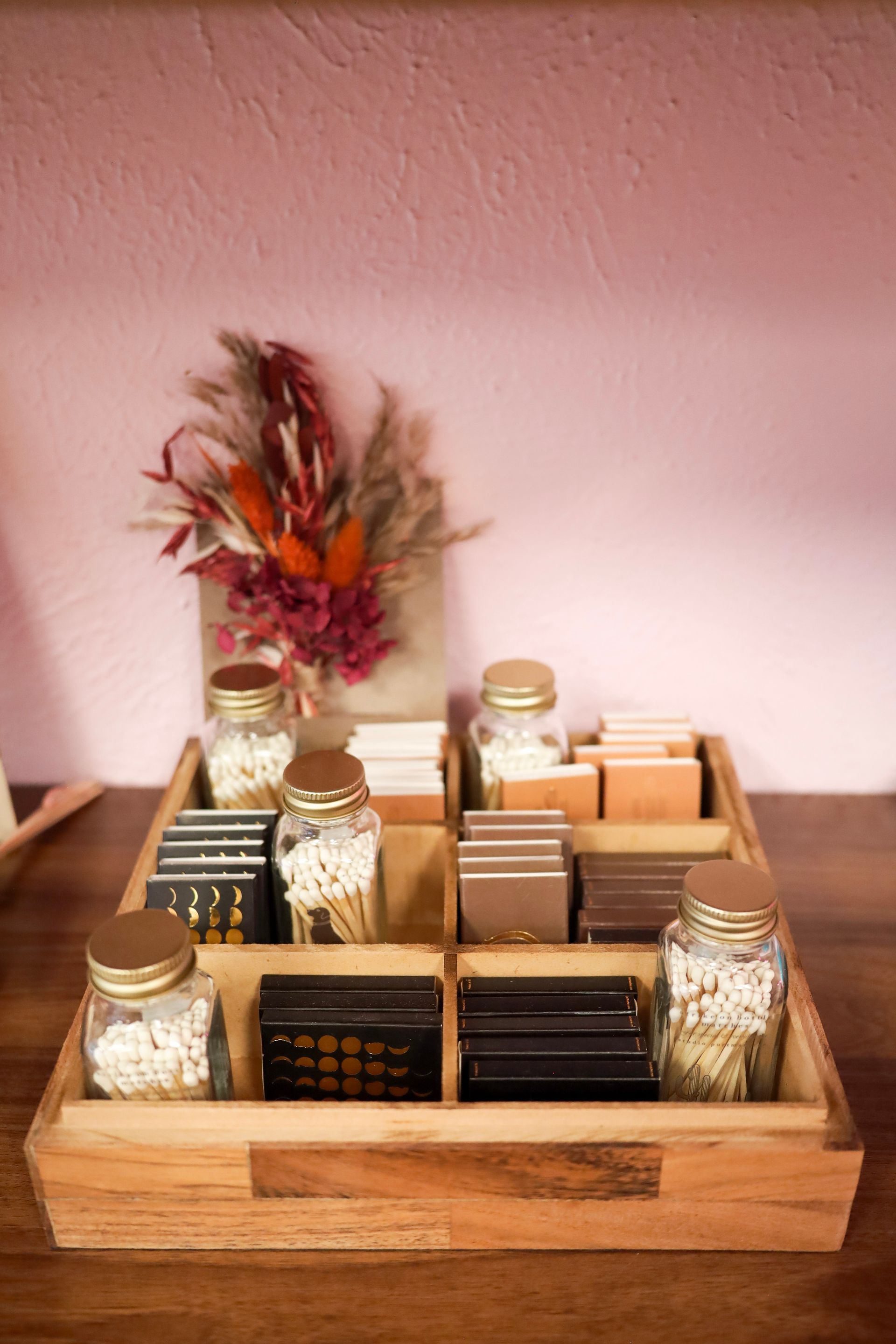 Wooden tray organizer with jars, boxes, and dried flowers on a wooden table against a pink wall