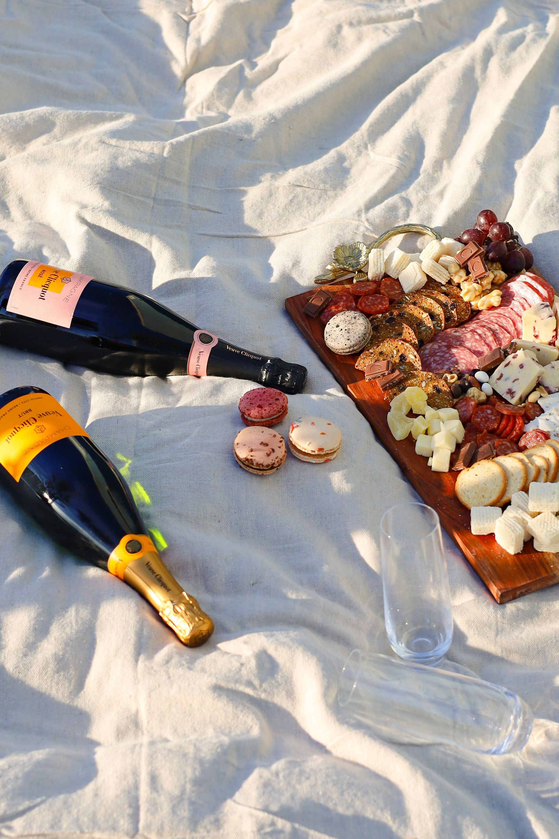Champagne bottles and a charcuterie board on a white blanket at a picnic