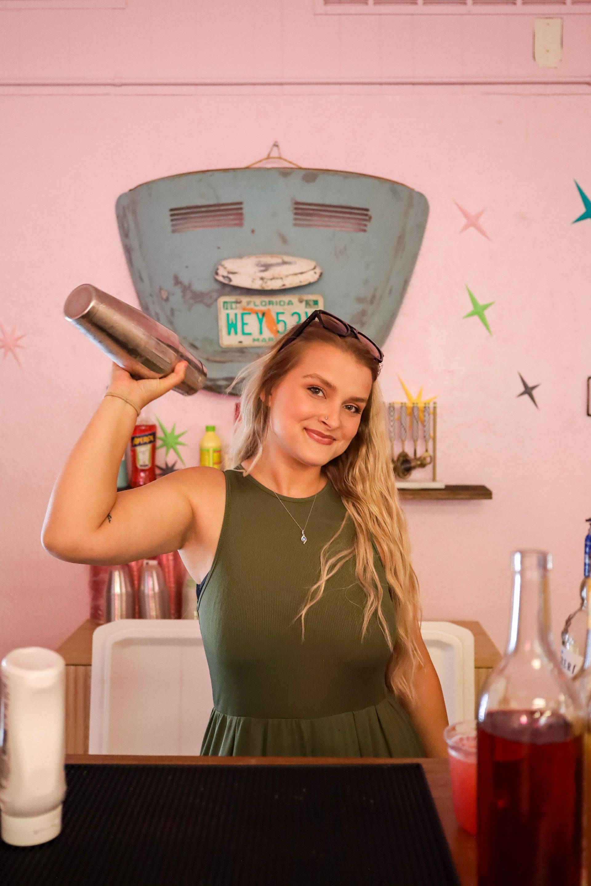 Person shaking a cocktail in a pink bar, with bottles and a vintage sign in the background