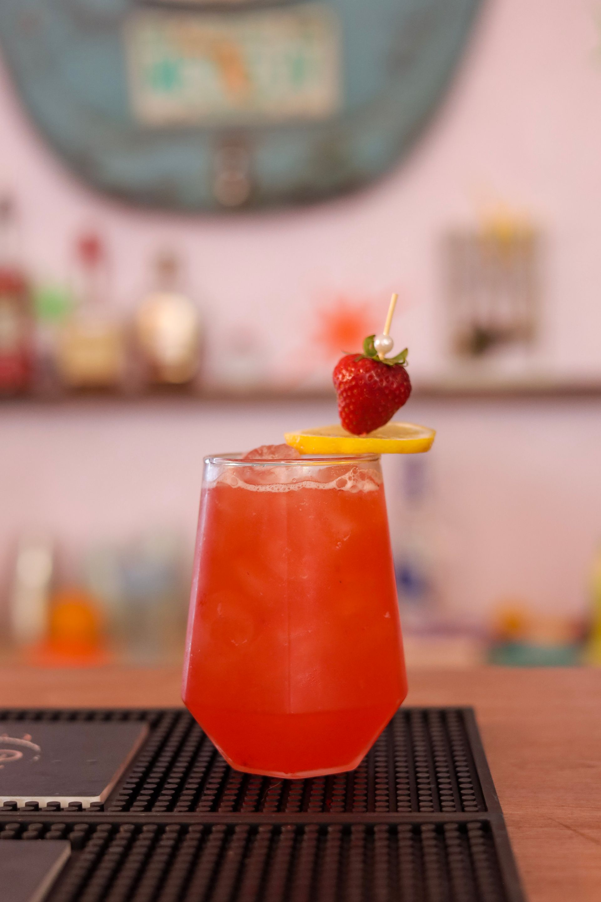 Red cocktail with a strawberry and orange slice garnish on a bar counter with a blurred café background