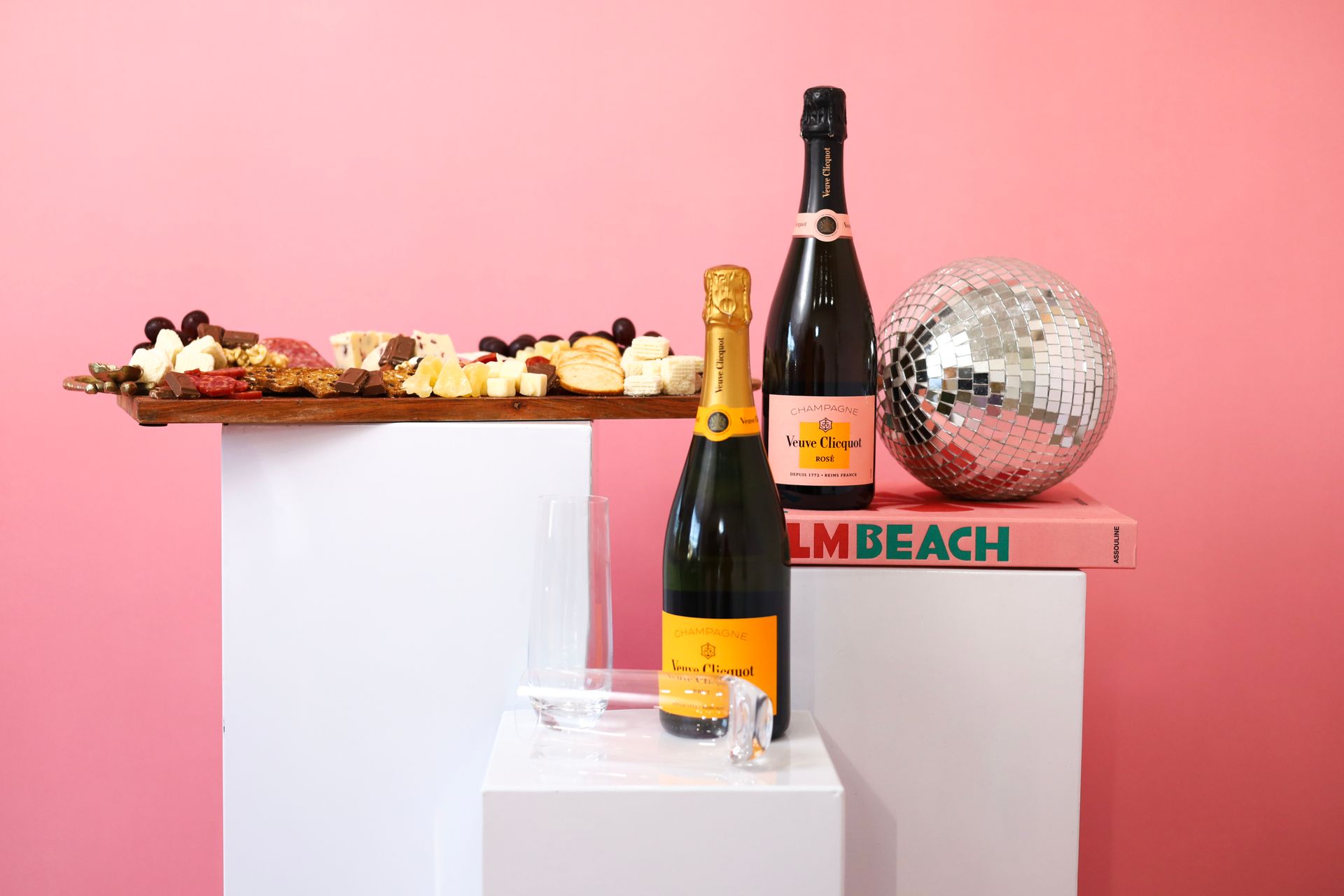Champagne bottles and a disco ball on white pedestals against a pink backdrop